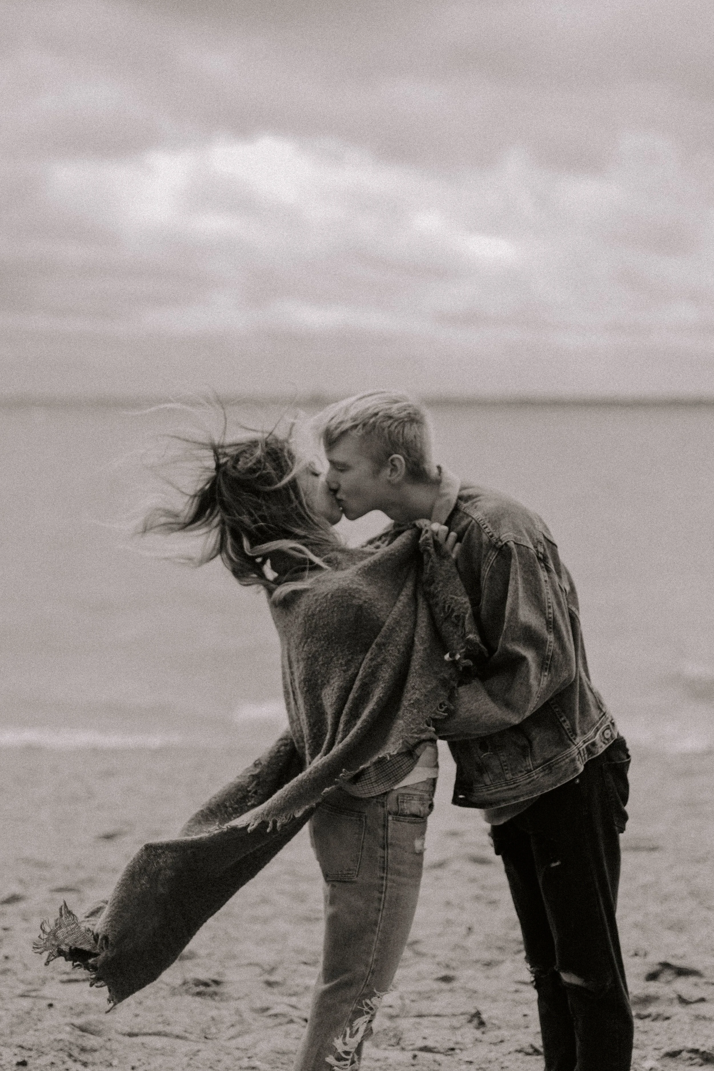 A young man and woman are kissing on a beach, with the woman wrapped in a large scarf and both wearing denim jackets. Their hair is blowing in the wind, and the ocean and cloudy sky are in the background.