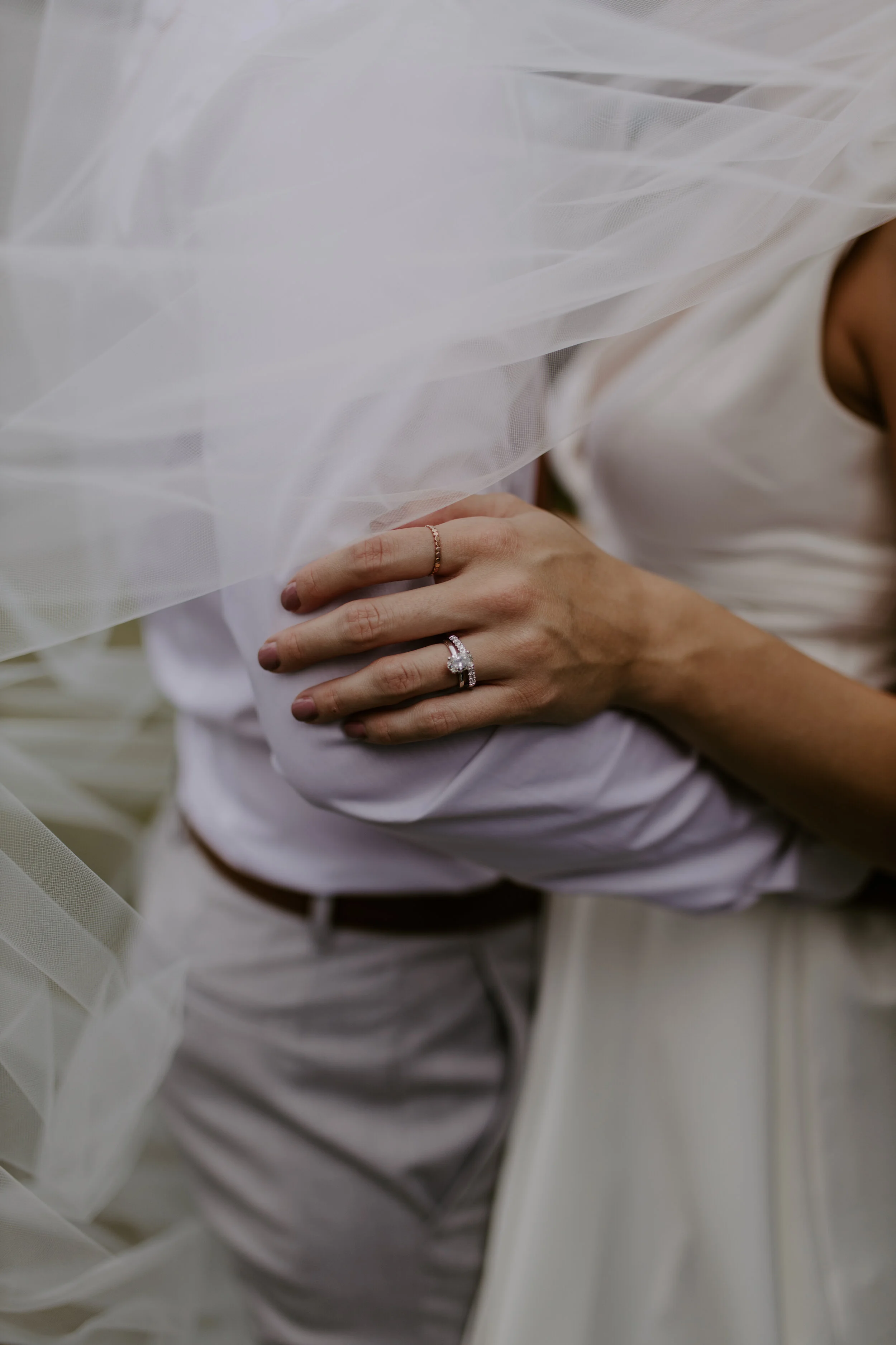 Person wearing a white dress featuring a sheer overlay, with rings on fingers, holding their arm.