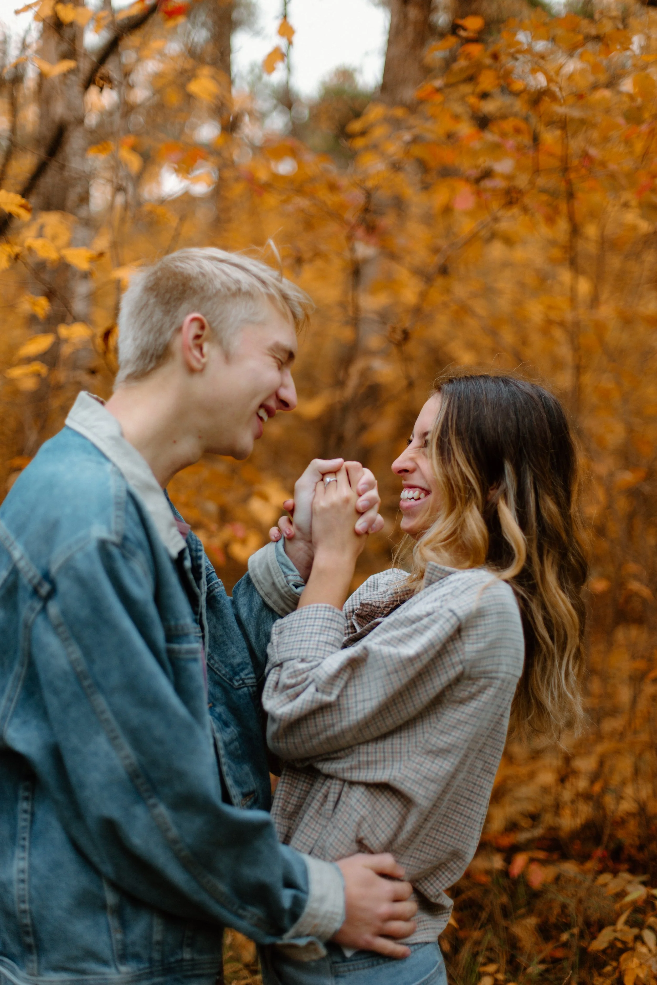 A young man and woman are smiling and holding hands in a forest with autumn leaves, leaning towards each other affectionately.