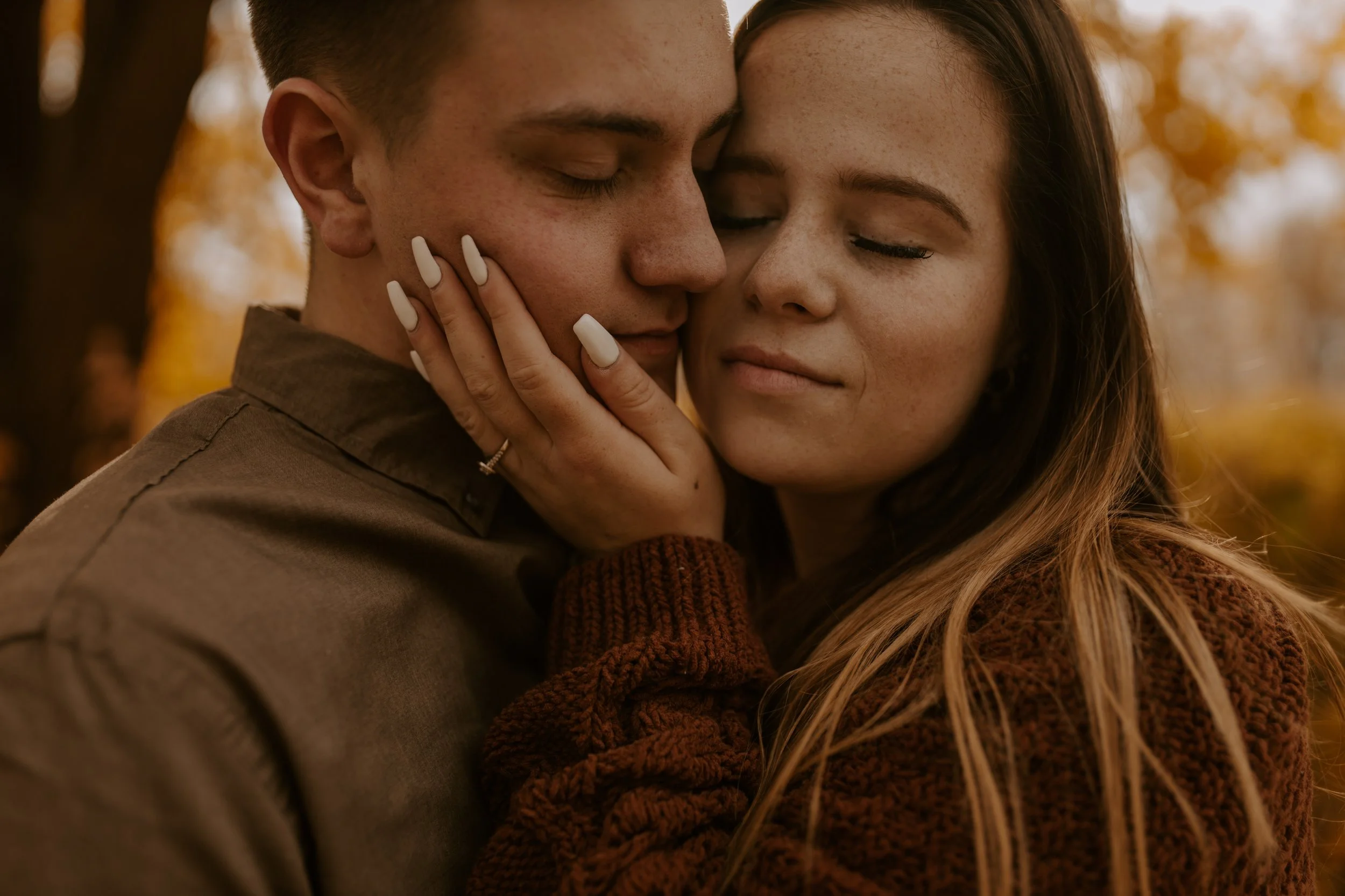 A close-up of a young couple with light skin and freckles embracing outdoors during fall, with warm-colored blurred trees in the background. The woman has long, wavy hair and is gently holding the man's face while both have eyes closed.