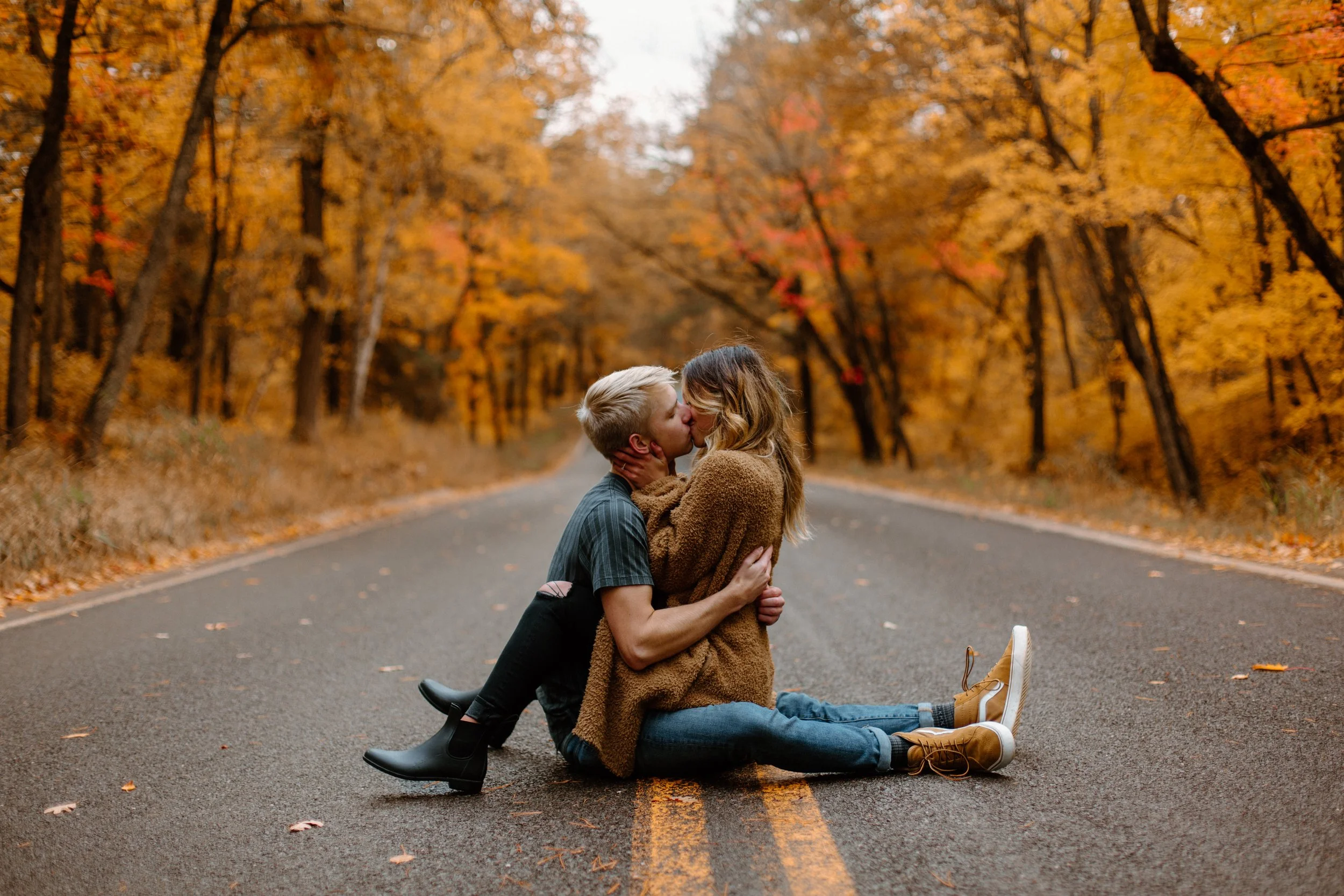 A couple is kissing on an empty road surrounded by fall foliage with orange and yellow leaves. The man is kneeling, and the woman is sitting on the ground, both smiling and dressed in cozy fall clothing.