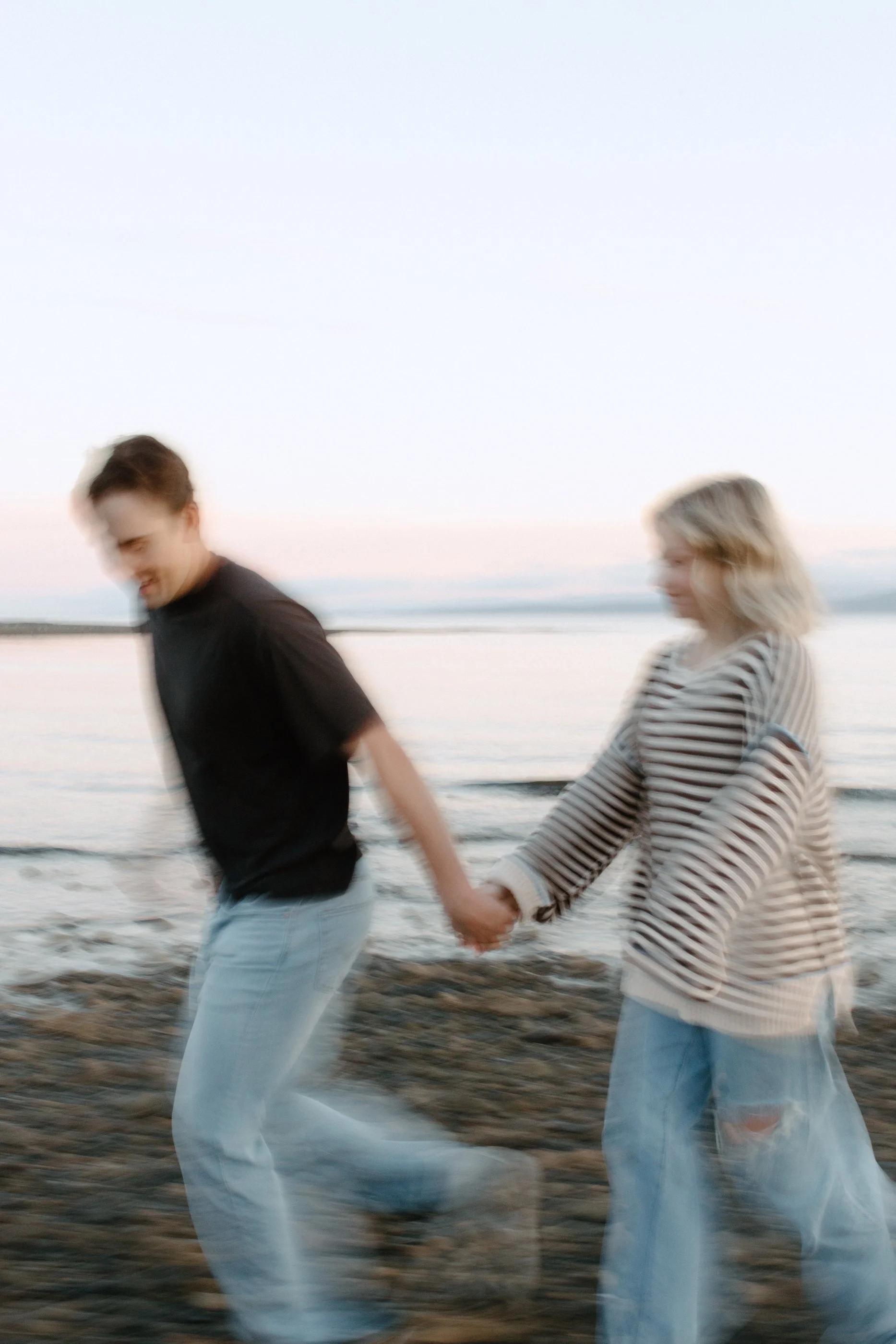 Two people, a man and a woman, holding hands and walking along a beach at sunset. The man is wearing a black t-shirt and light blue jeans, and the woman is wearing a striped long-sleeve shirt and ripped jeans. The background features the ocean with g