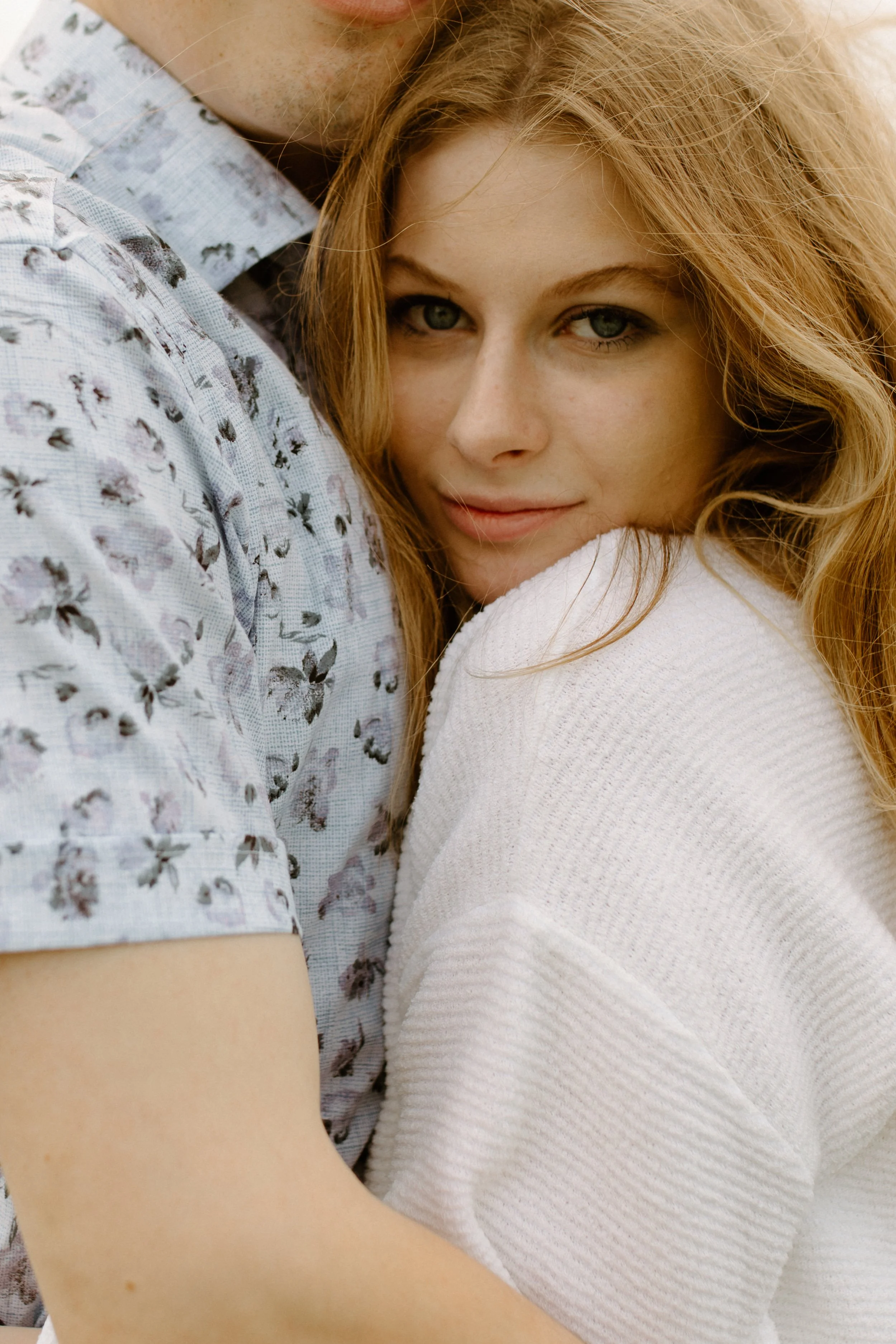 Close-up of a young woman with red hair and gray eyes hugging a man, with part of his face and shoulder visible, wearing a light blue floral shirt. The woman has a slight smile and rests her head on his shoulder.