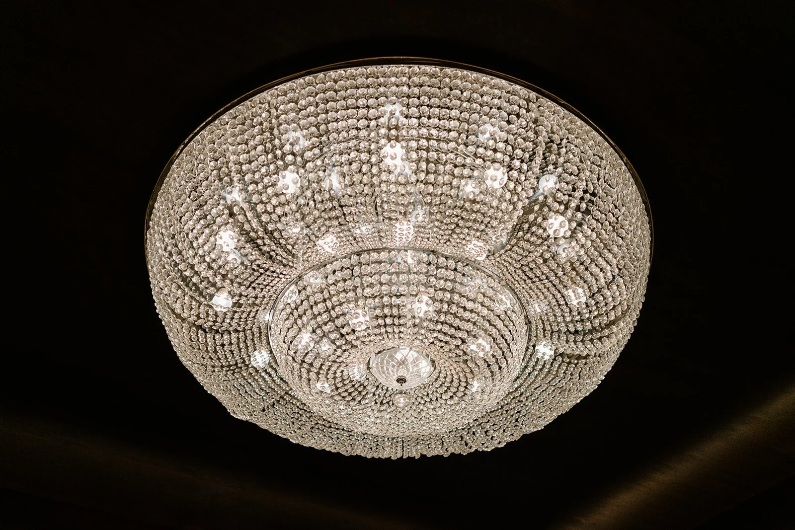 Close-up of a large, round crystal chandelier with multiple tiers, hanging from a dark ceiling.