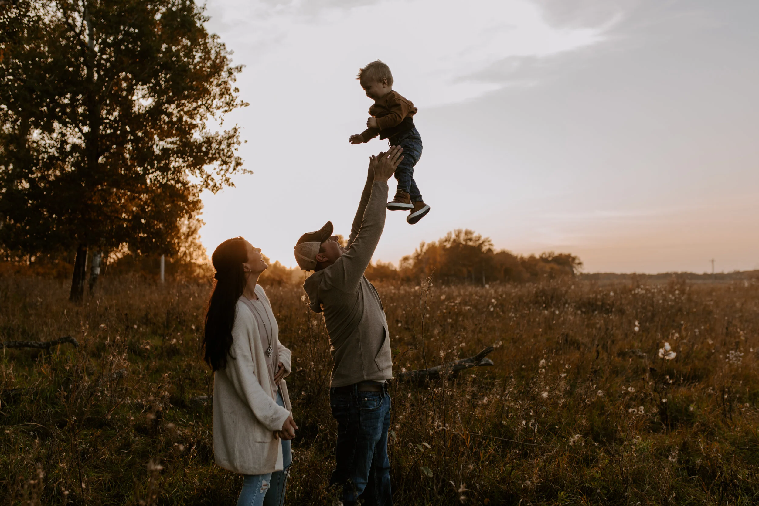 A family of three, two adults and one child, outdoors during sunset. The man is lifting the boy into the air, and the woman is standing nearby, watching, in an open field with trees in the background.