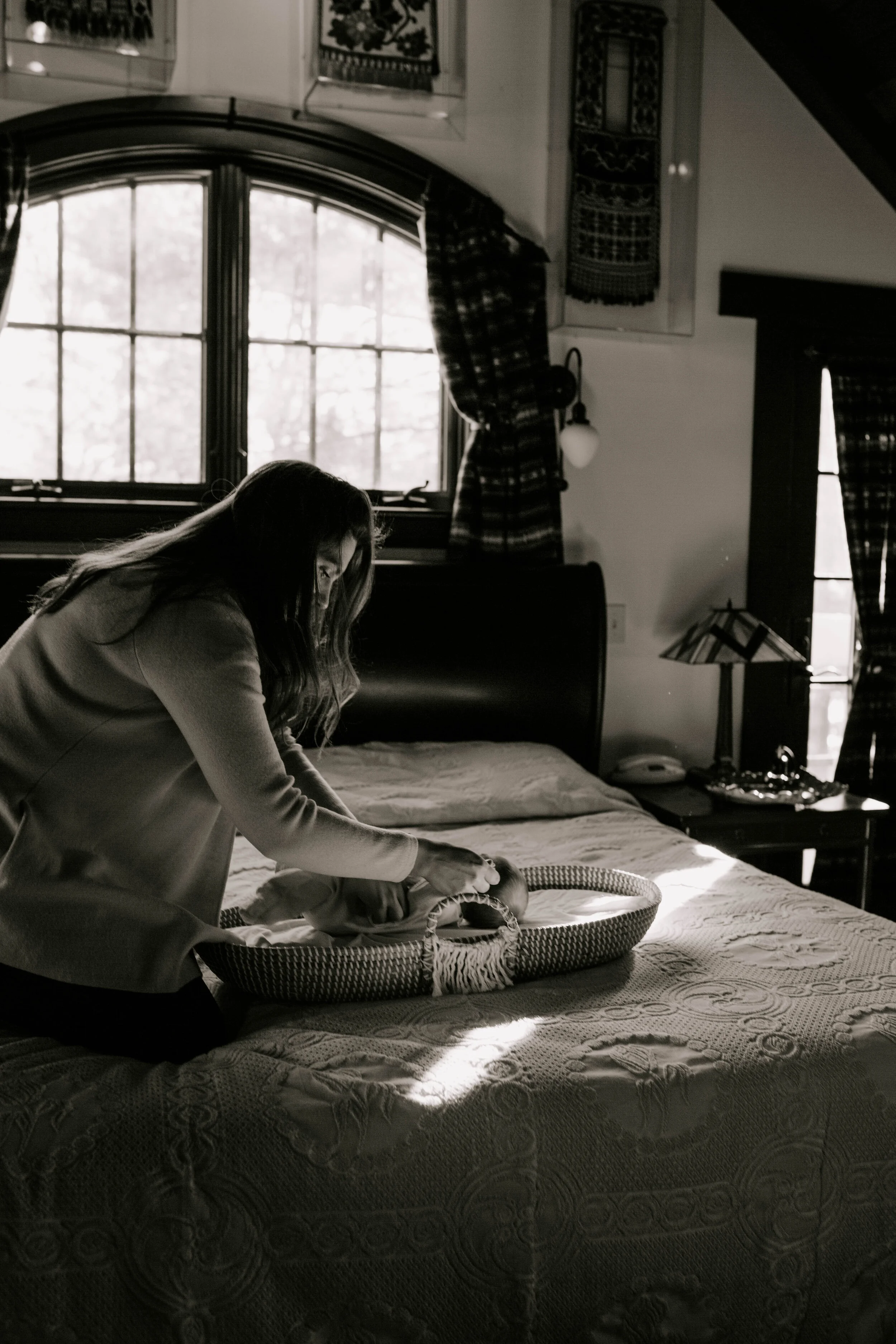A woman is leaning over a bed, possibly tending to a pet in a wicker basket, in a cozy bedroom with large window, curtains, and patterned quilt on the bed, in black and white.