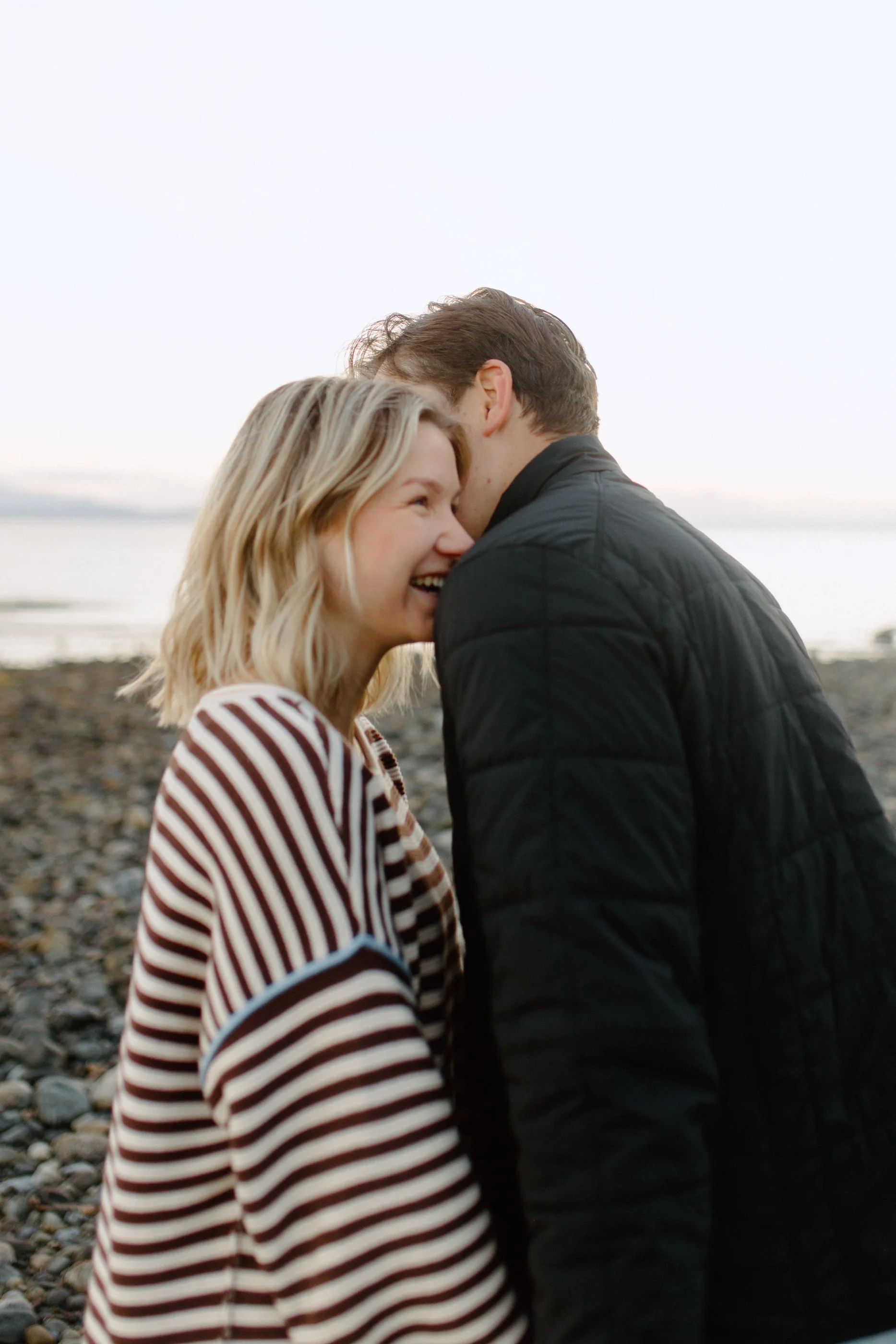 A smiling blonde woman and a man with brown hair are close together, smiling and leaning their foreheads and noses against each other on a beach at sunset, with the ocean in the background.