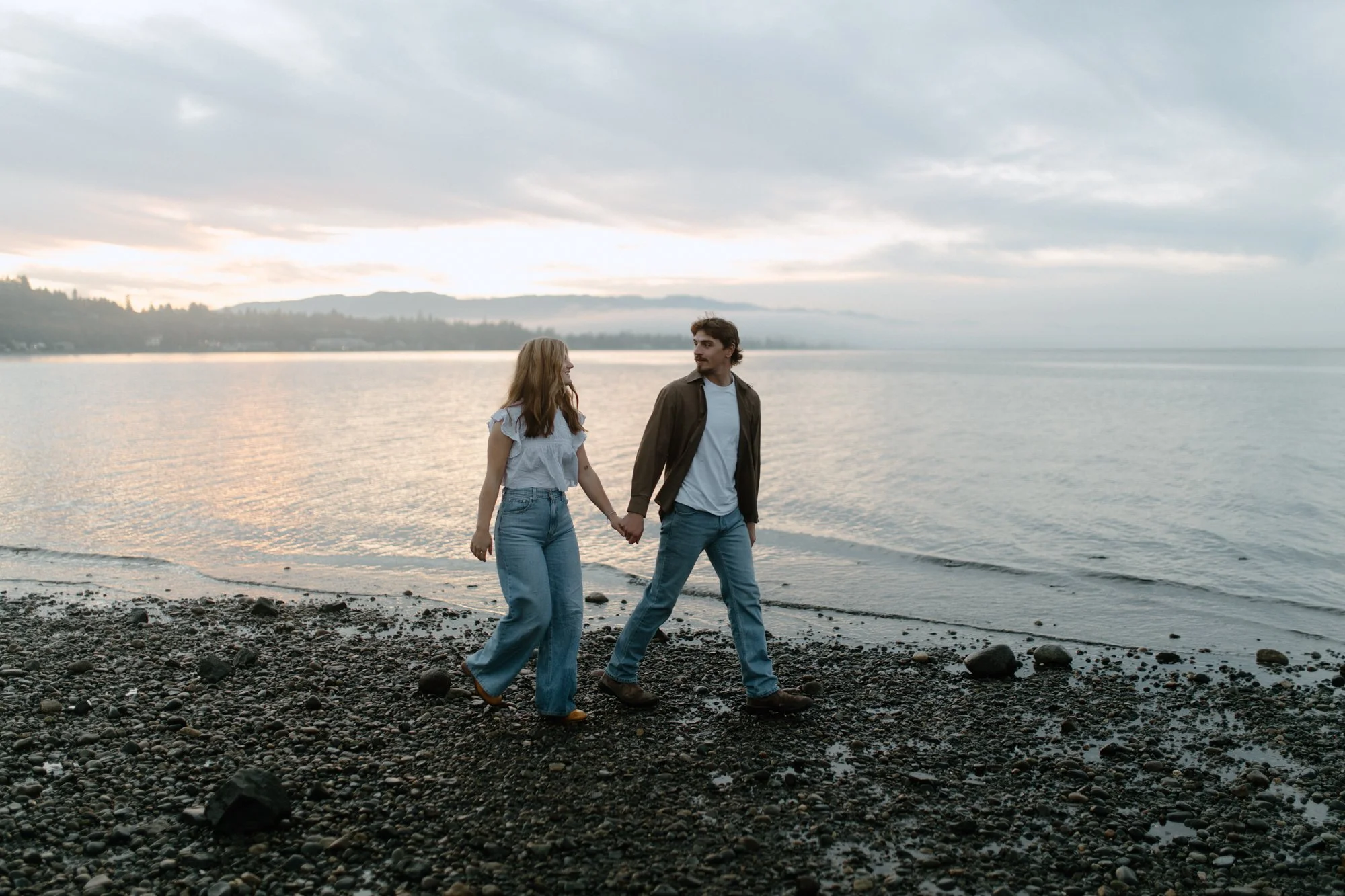 A young couple holds hands while walking along a pebbled beach at sunset, with calm water and mountains in the background.