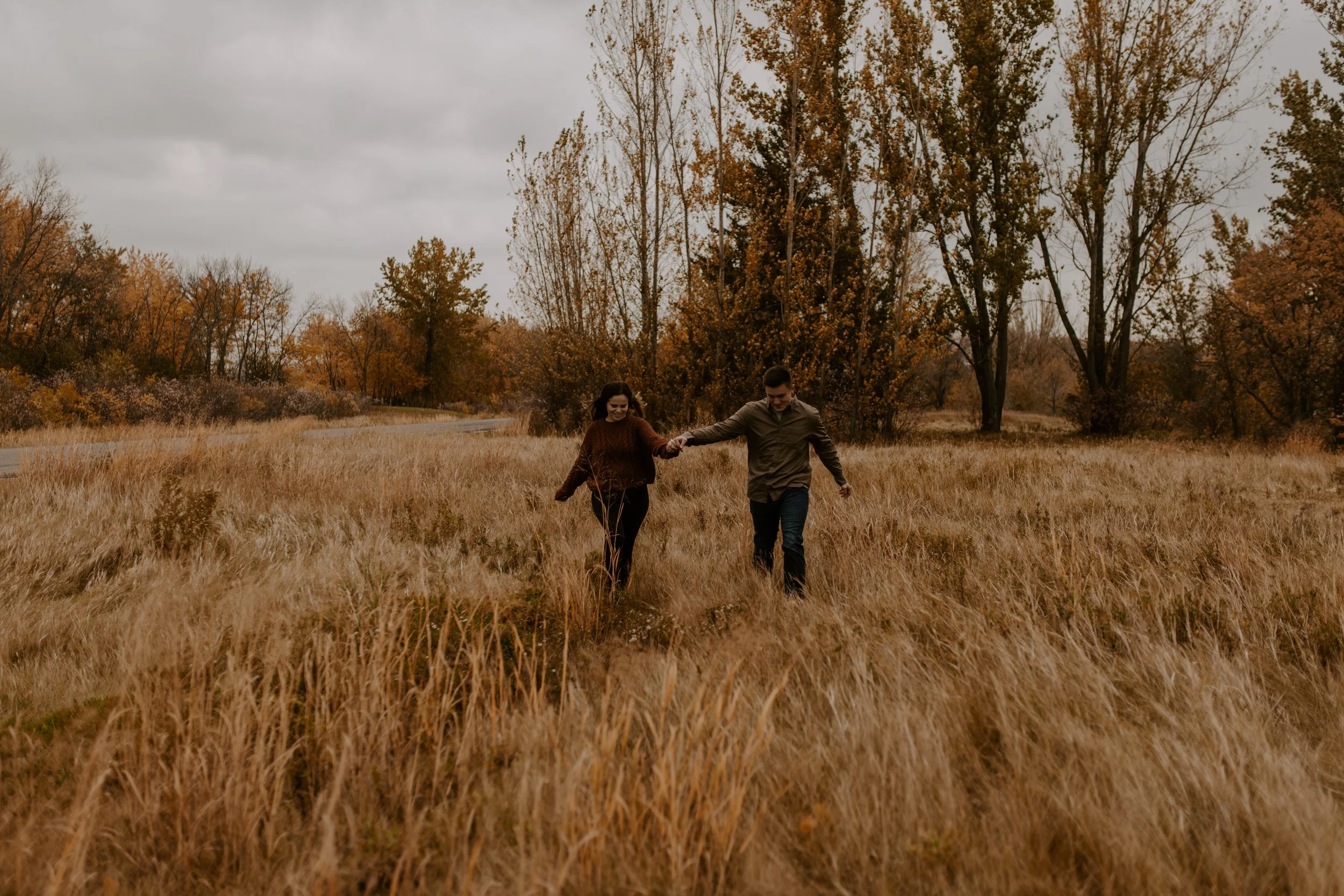 A young man and woman walking hand in hand through a field with tall, golden grass during autumn, surrounded by trees with orange and yellow leaves under a cloudy sky.