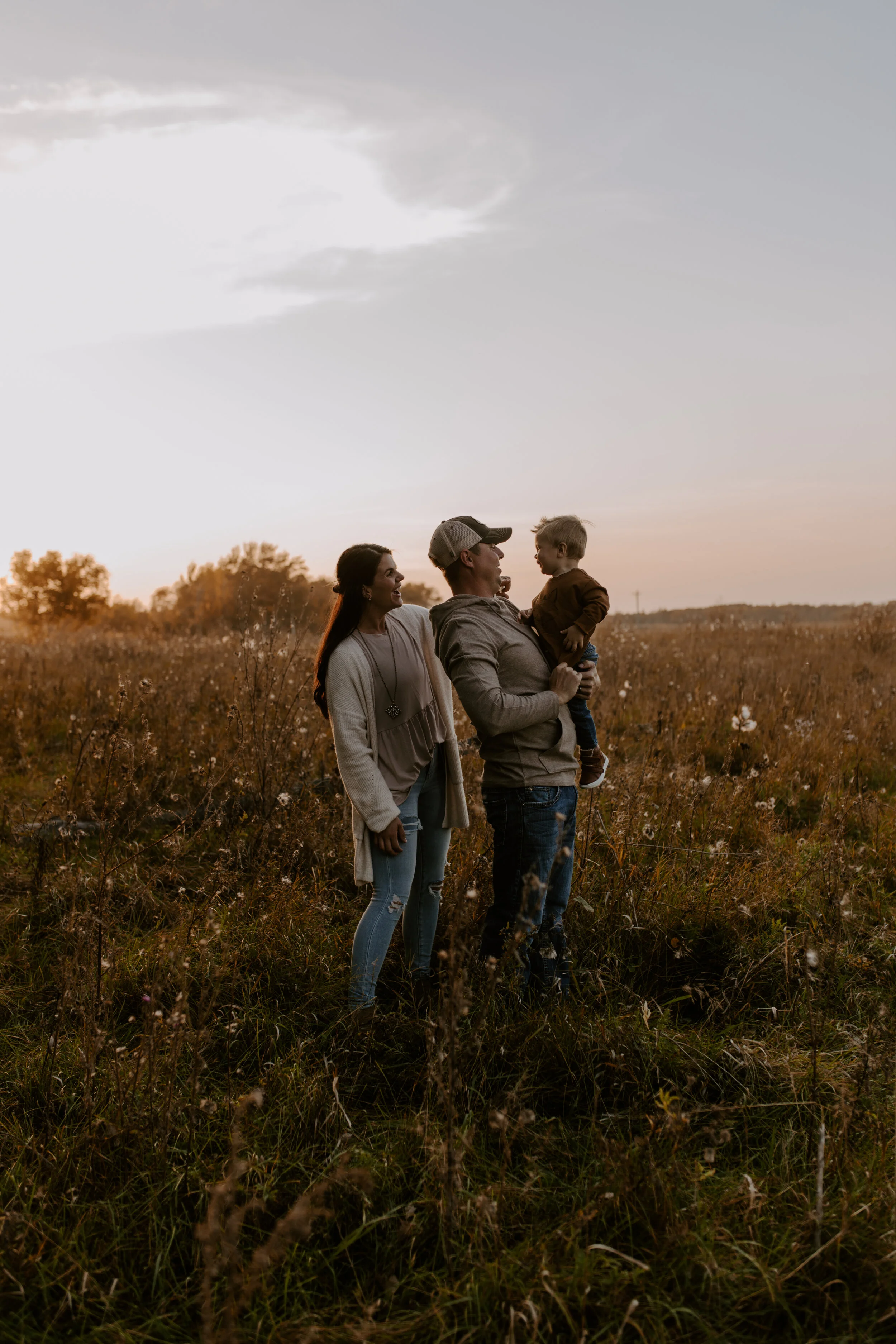 Family of three standing in a field at sunset, with trees in the distance and a partly cloudy sky overhead.