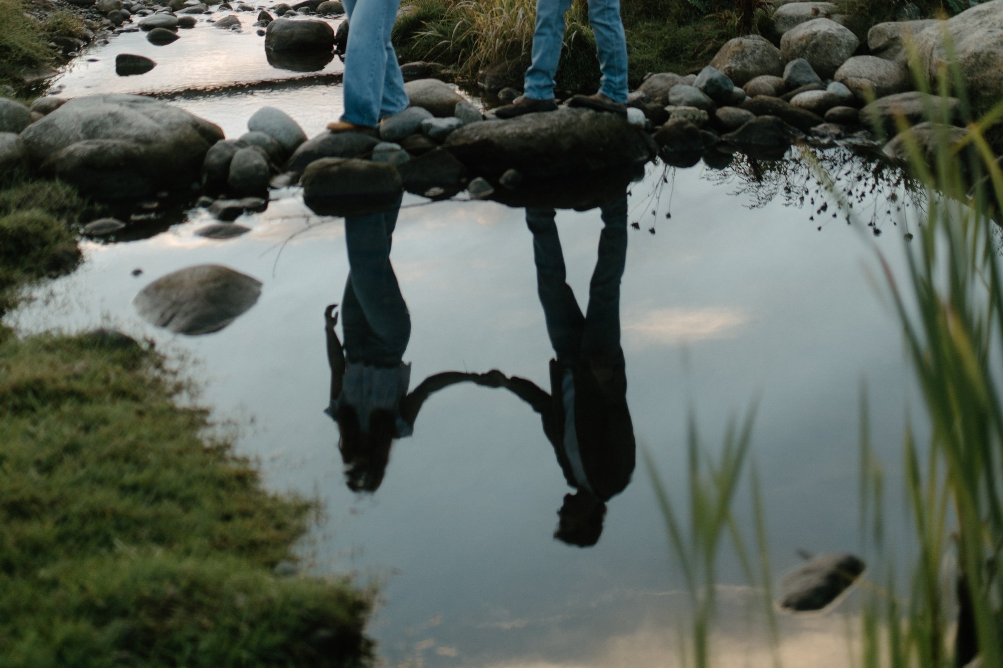 A person standing on rocks near a calm river, with their reflection visible in the water.