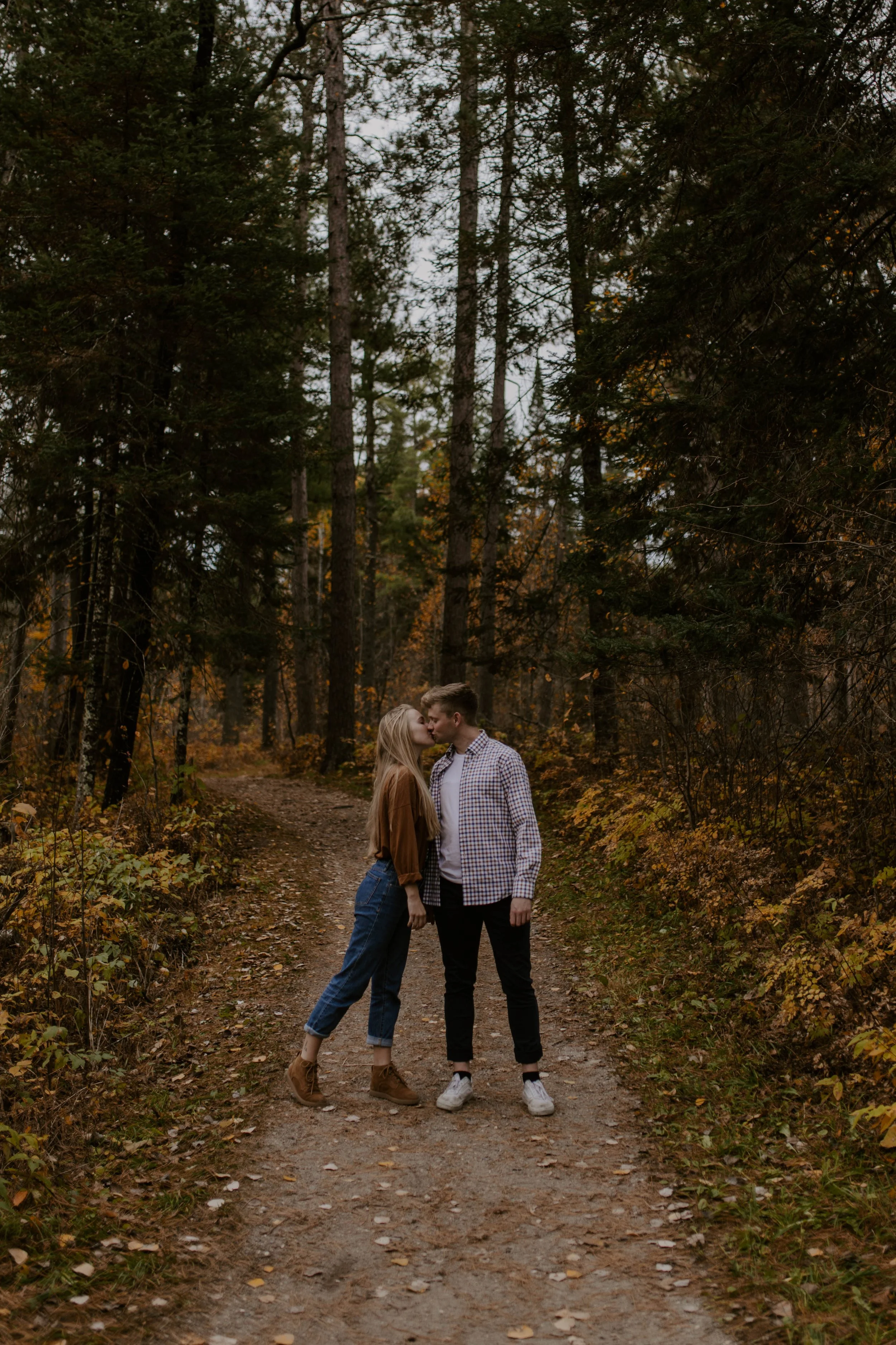 A young couple standing on a dirt path in a dense forest, leaning in for a kiss with trees and fall foliage around them.