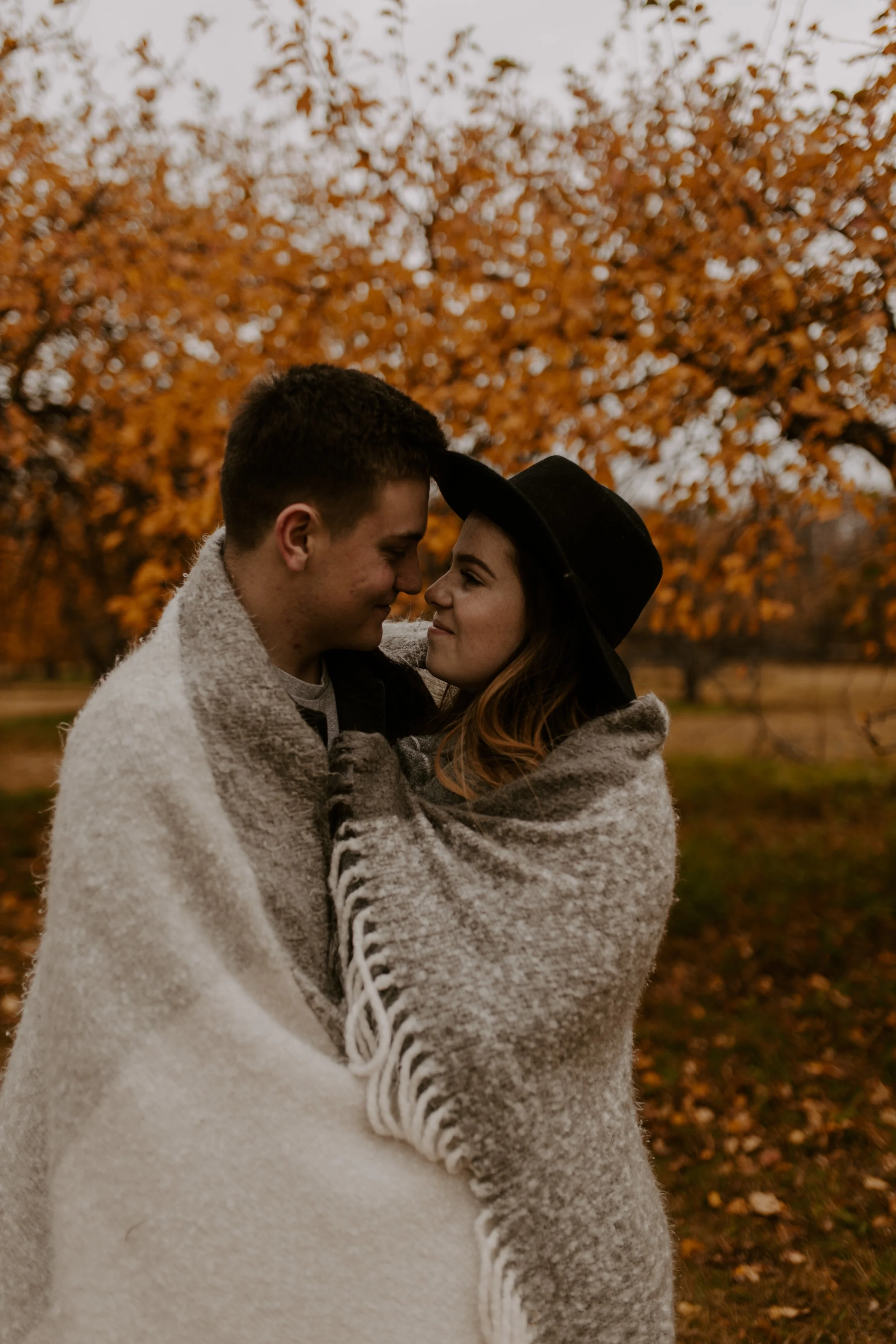 A couple stands close together outdoors in autumn, embracing and smiling at each other with their foreheads touching, surrounded by orange-leaved trees.