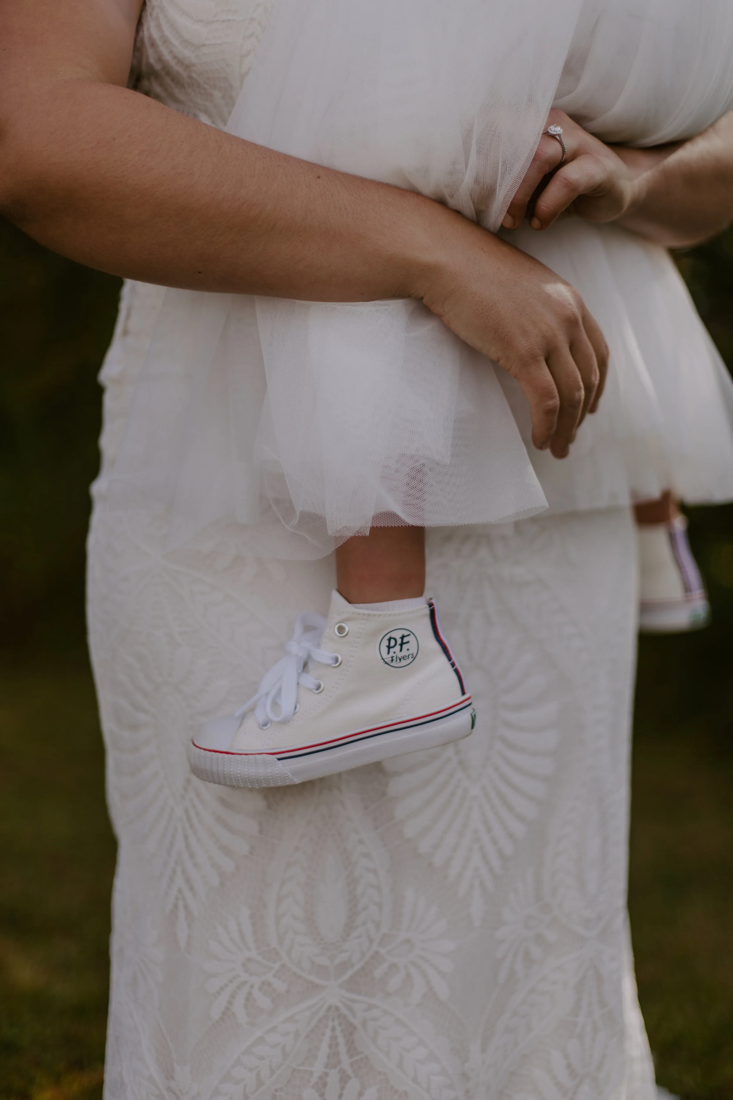 Close-up of a person's arm and a small child hanging from the person's hand, wearing white sneakers with striped soles, in front of a white lace dress.