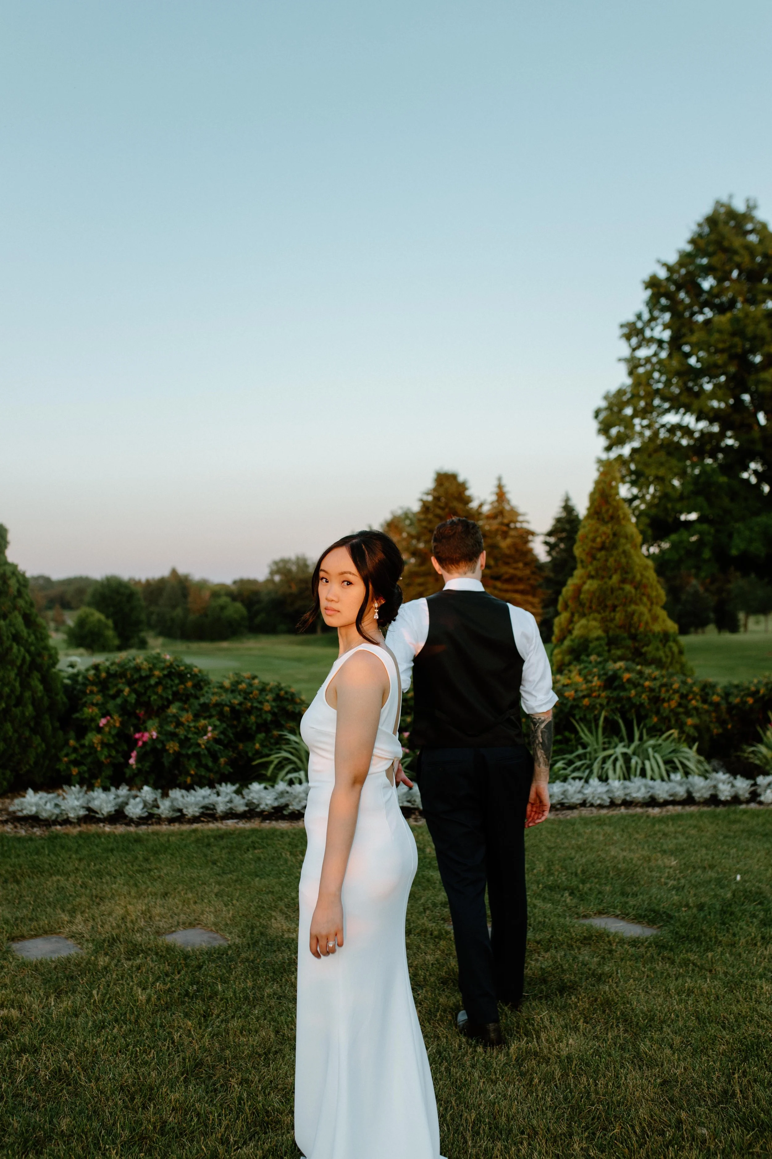 A woman in a white dress standing in the foreground and a man in a vest and white shirt standing with his back turned among landscaped garden bushes and trees during the daytime.