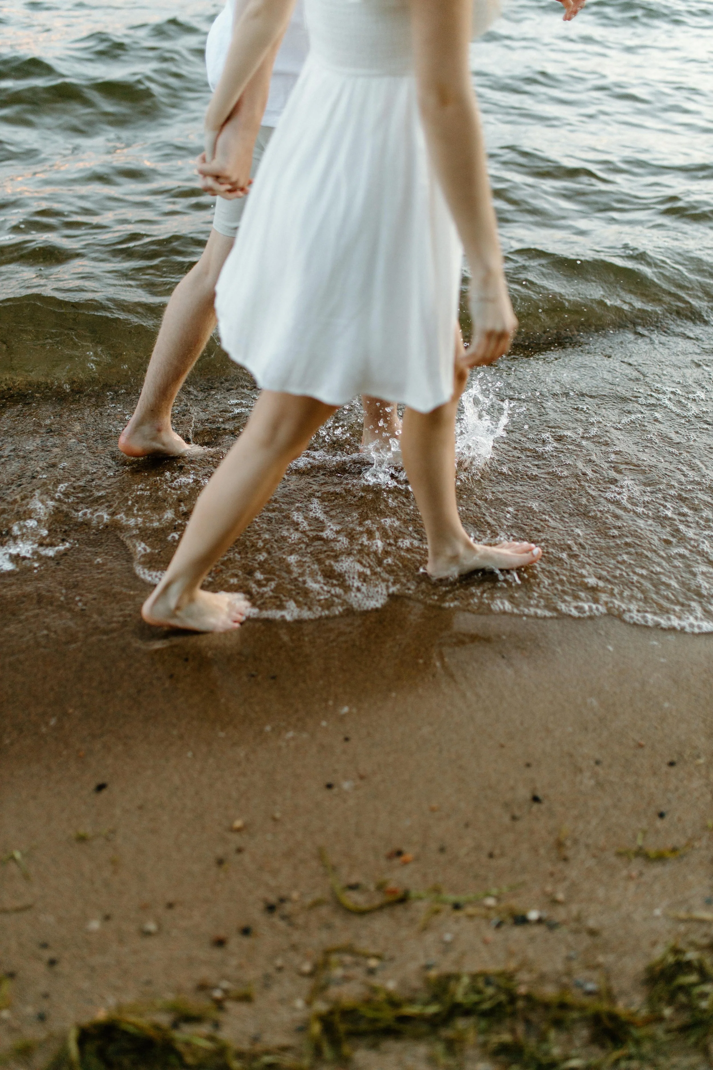 People walking barefoot along the shoreline, with waves touching their feet, wearing casual summer clothing.