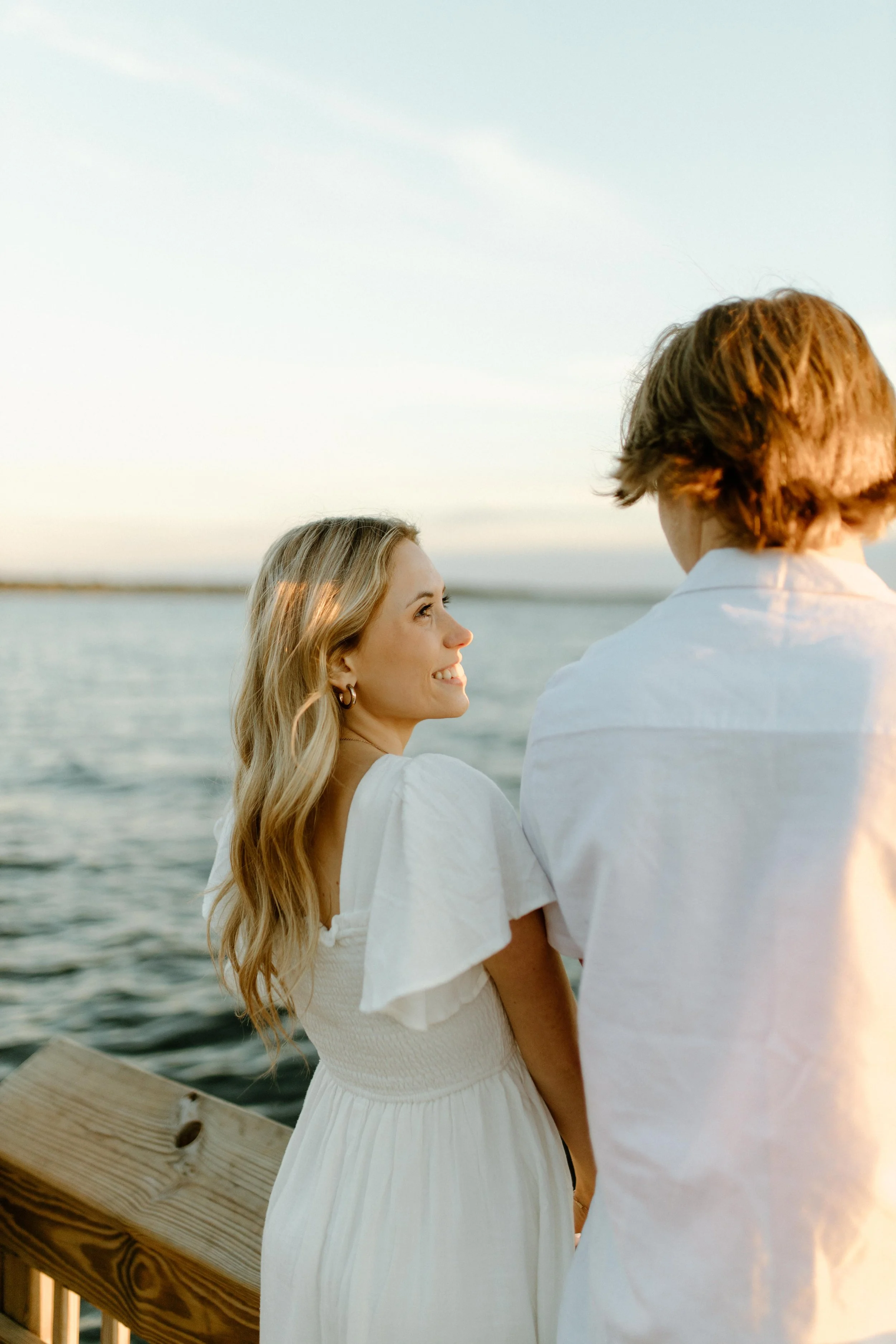 A woman with long blonde hair smiling and looking at a man with short red hair by the water, both wearing white clothing, during sunset.