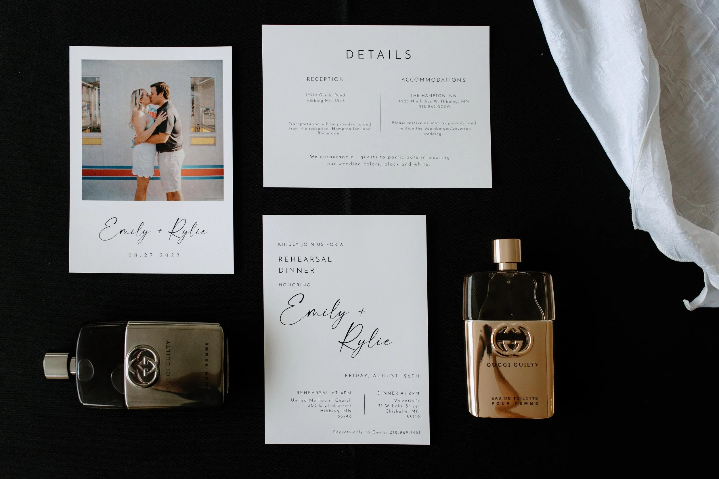 A wedding invitation suite with a photo of a couple kissing, wedding details, and perfume bottles on a black surface with white fabric draped on the right.