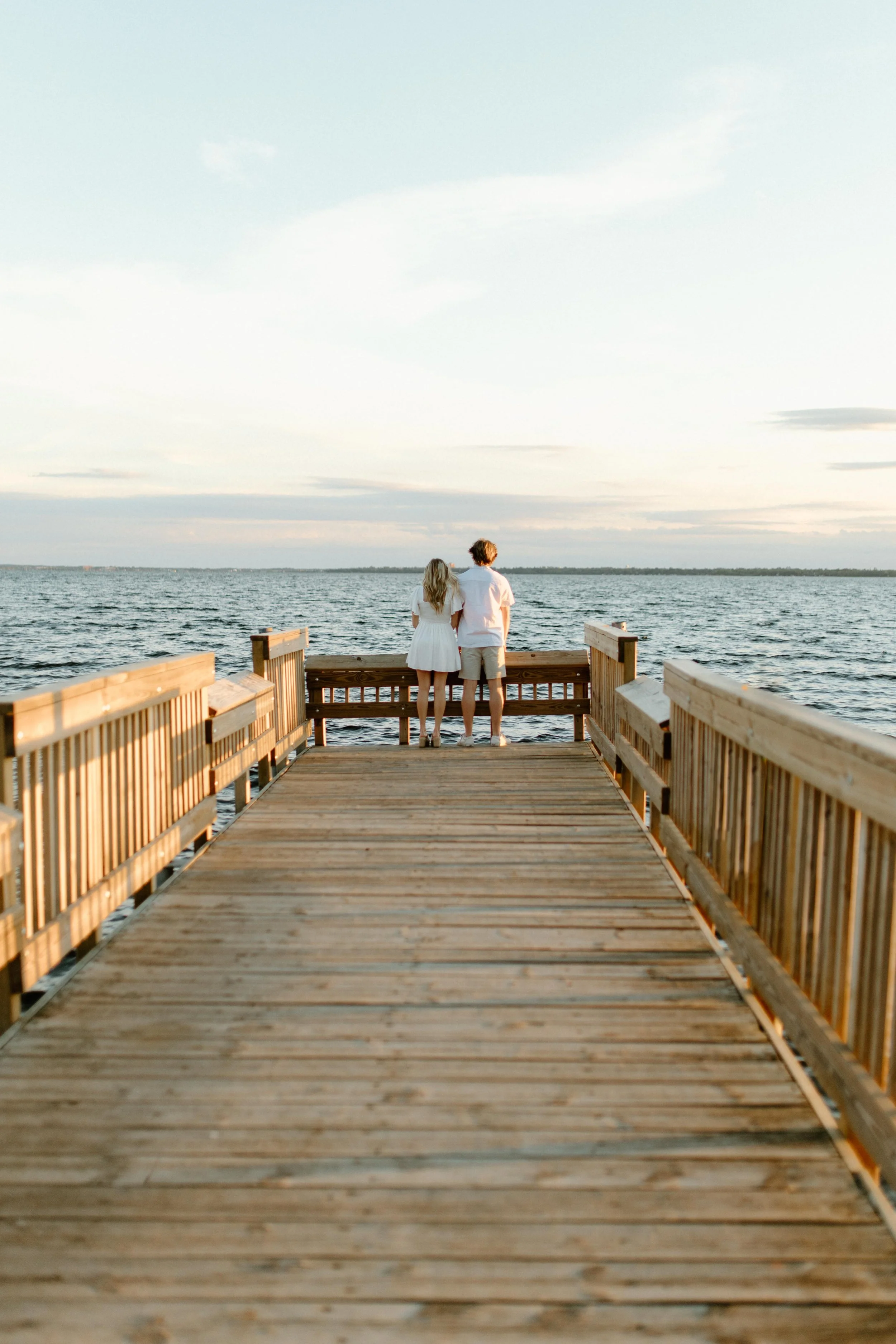 A couple standing on a wooden pier by the water, facing away, watching the horizon at sunset.