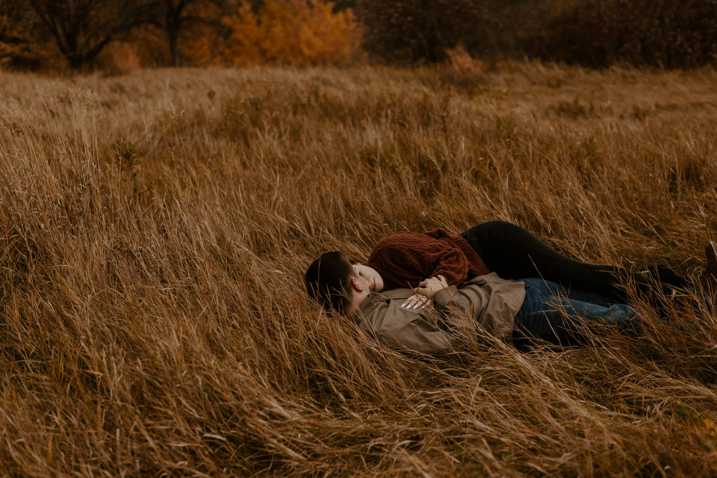 A couple lying on the grass in a field during autumn, embracing each other with their faces near each other's.