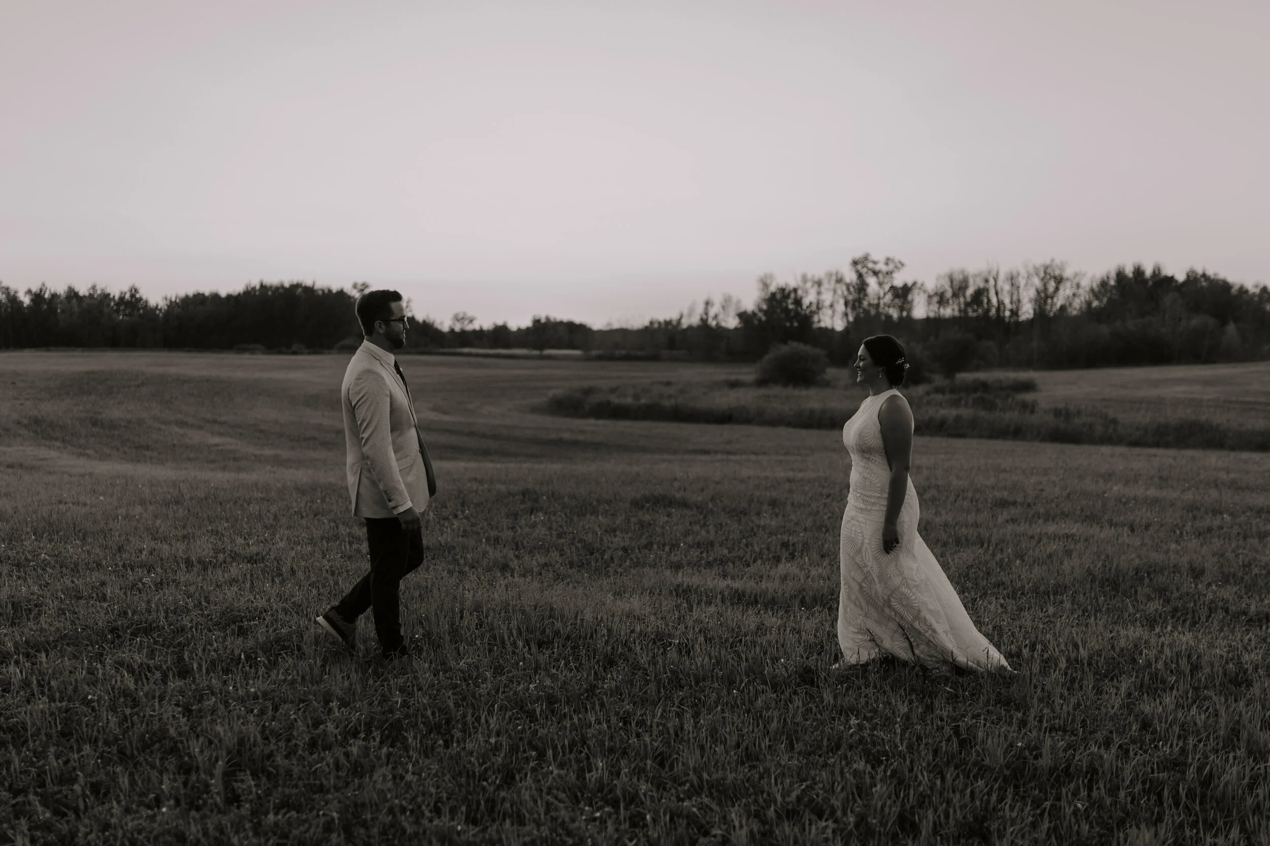 A black-and-white photo of a man and woman standing facing each other in an open field, with trees in the background, during sunset or dusk.