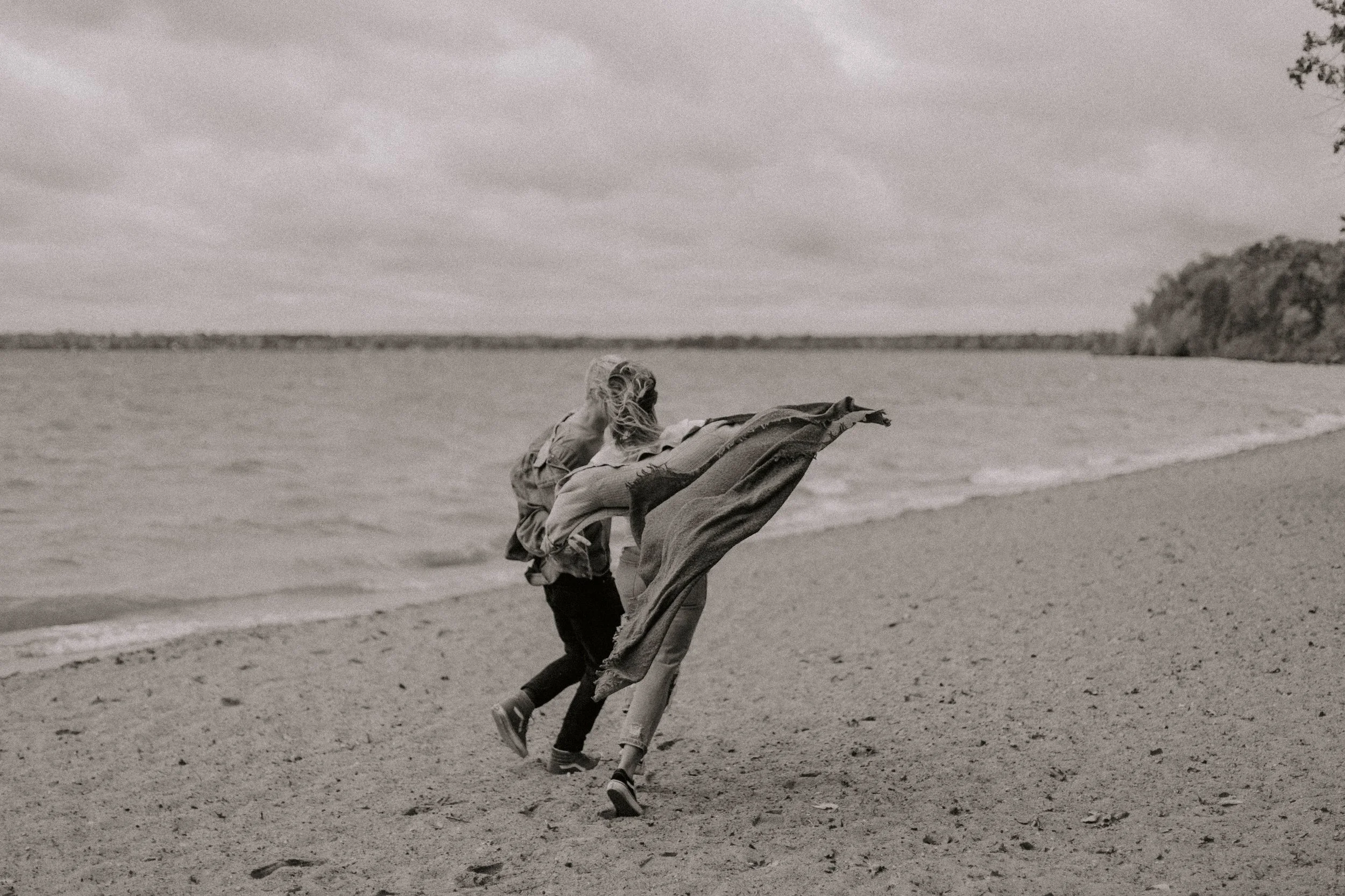Two children running along the shoreline at the beach, with a cloudy sky and trees in the distance.