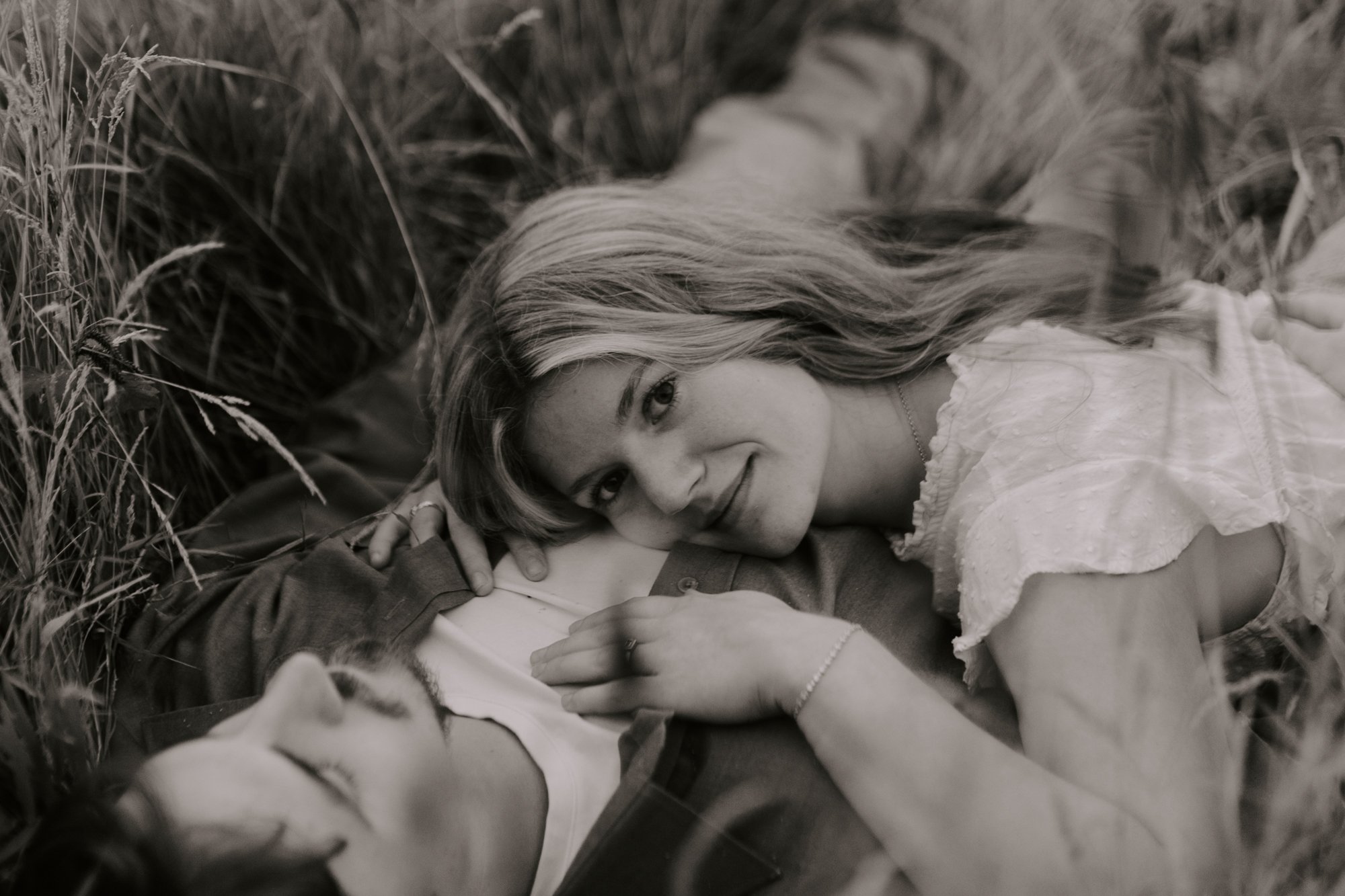 A woman with long wavy hair lying on the ground outdoors in tall grass, looking up and smiling. She is wearing a light-colored dress.