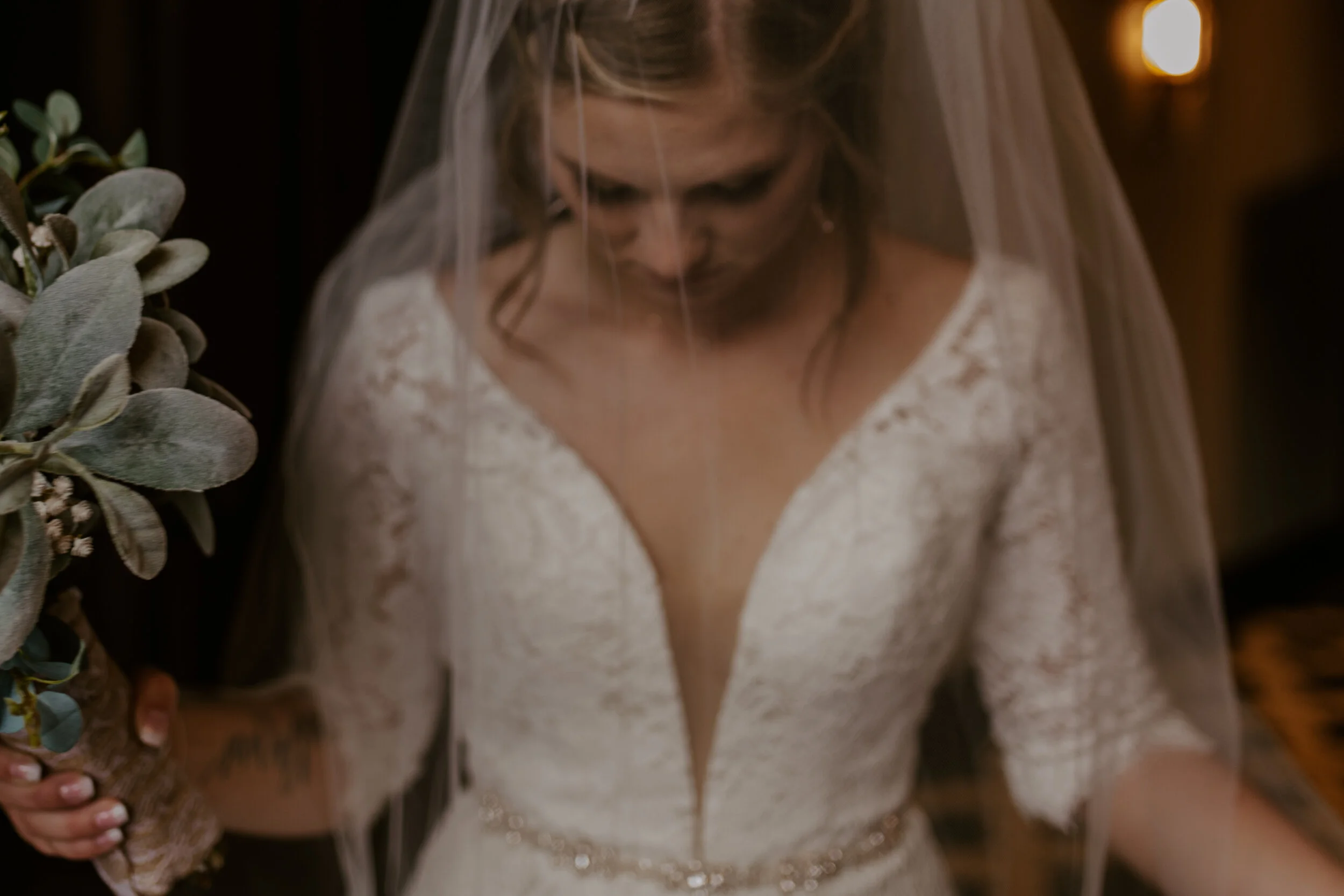 A bride in a white lace wedding dress with a veil, looking down, holding a bouquet, standing beside another person in a patterned outfit.