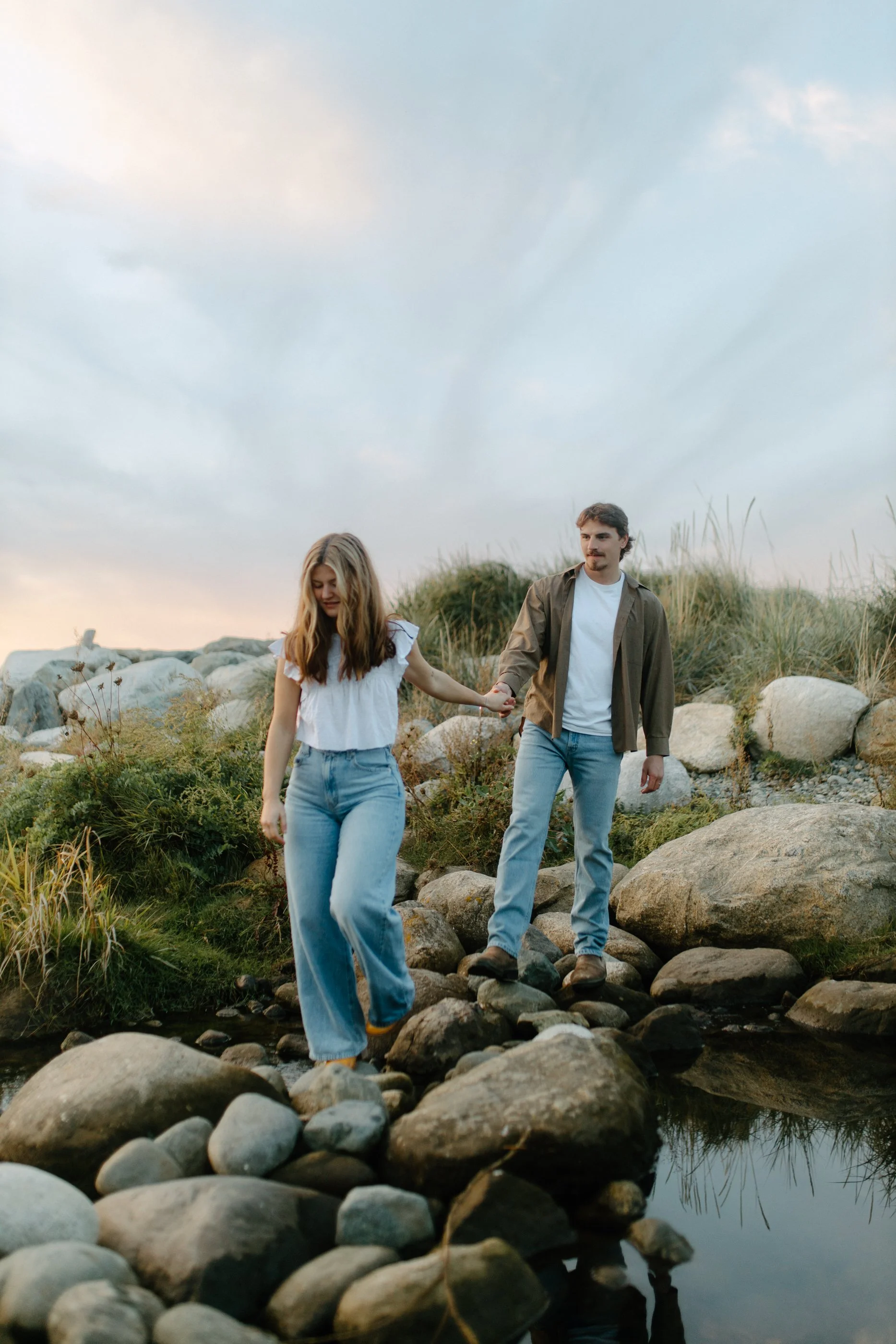 A couple walking across rocks along a small stream or pond outdoors, with tall grass and a cloudy sky in the background.