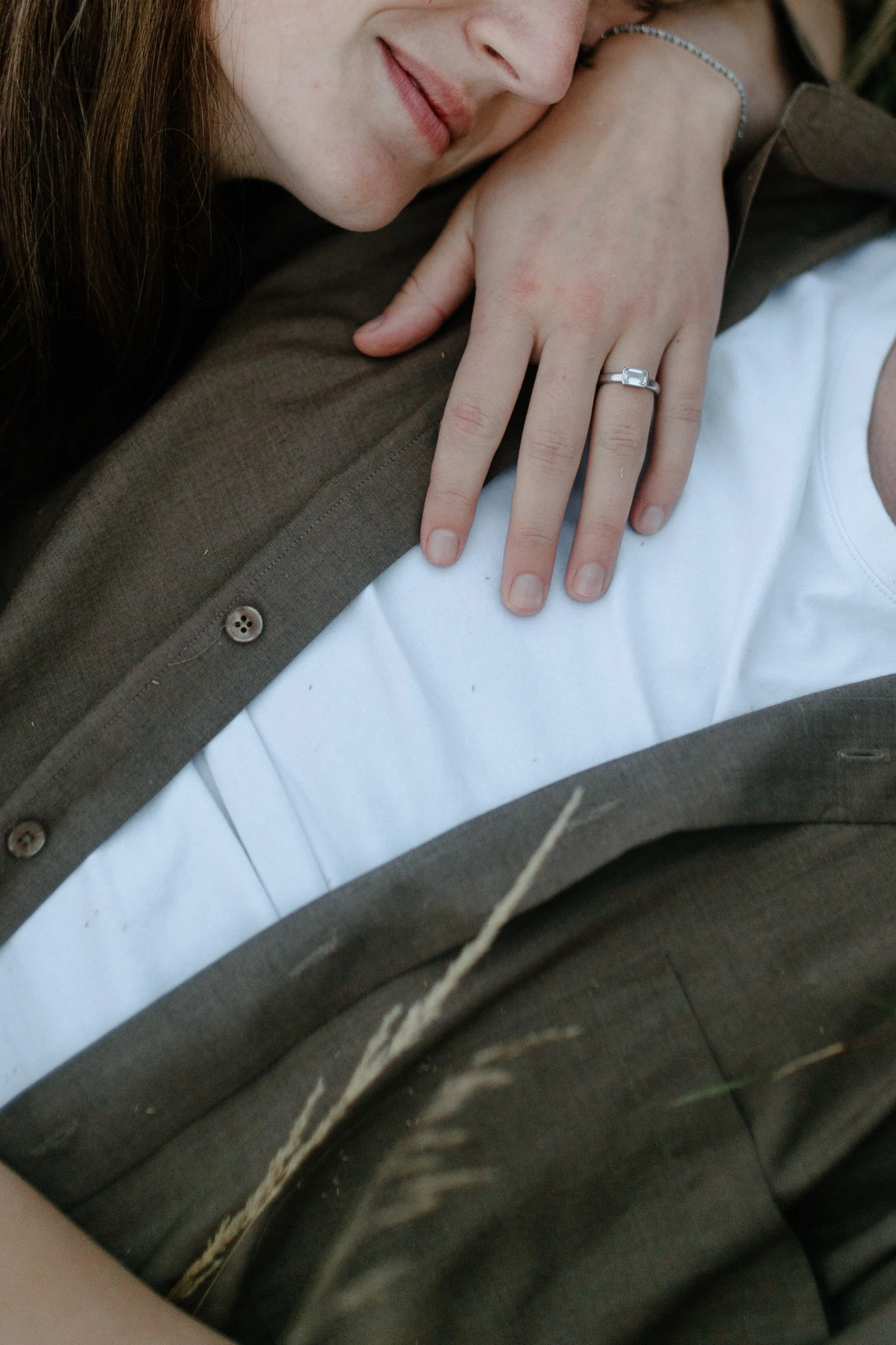 Close-up of a woman's face resting on her hand, wearing a diamond ring and a bracelet, with a brown shirt/coat and white shirt visible.