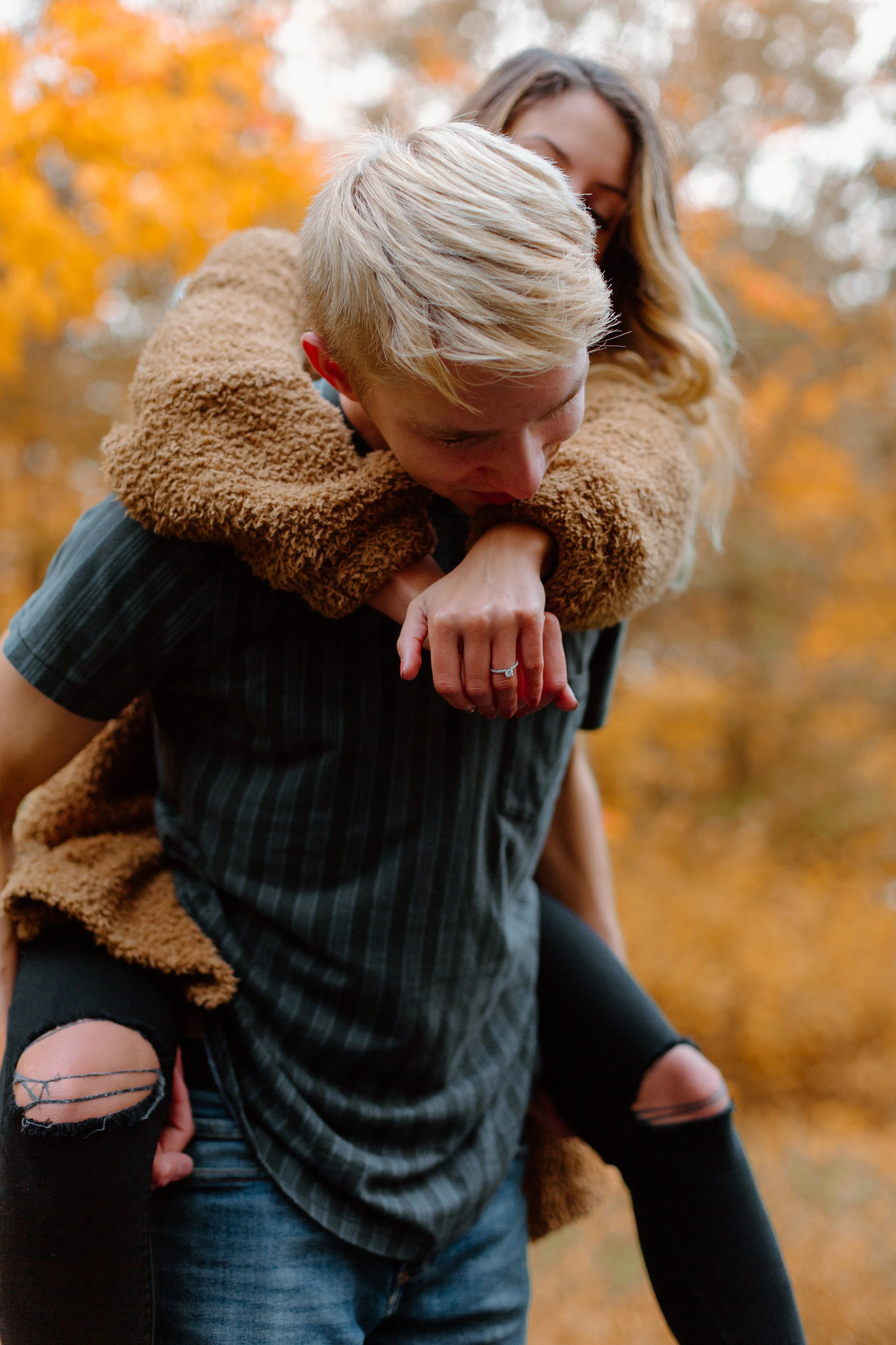 A young man giving a piggyback ride to a young woman in an outdoor fall setting with orange and yellow leaves.
