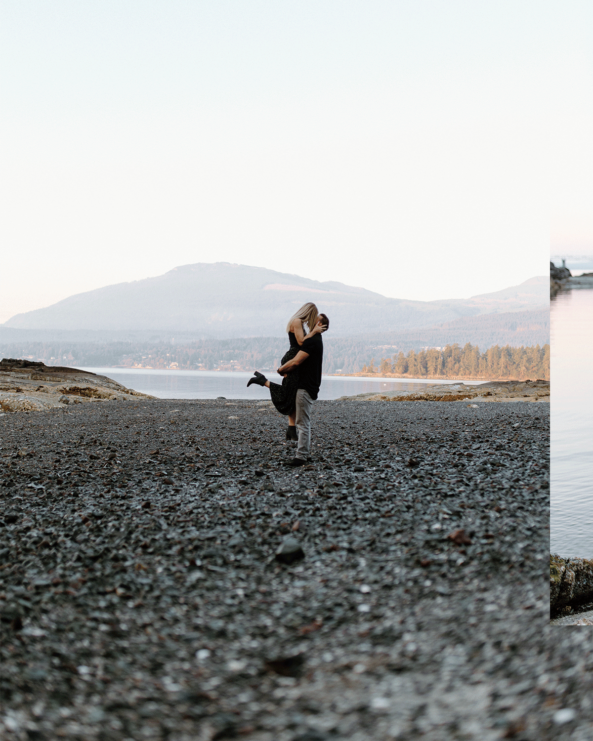 A couple on a rocky beach with mountains and a lake in the background, the man lifting the woman as they embrace.