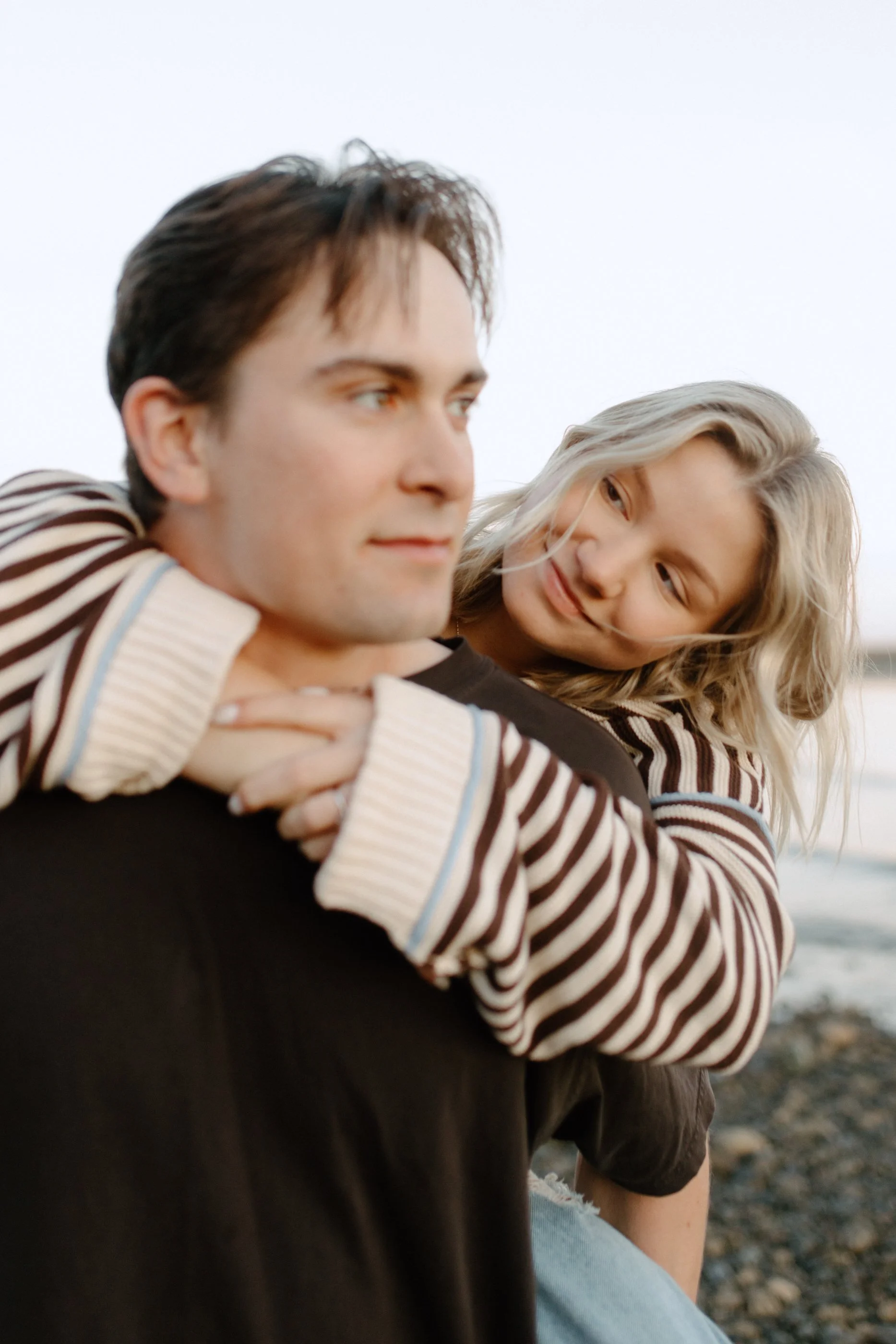 A young man and woman sitting together outdoors near a body of water, with the woman resting her head on the man's shoulder, both looking at each other with gentle expressions.