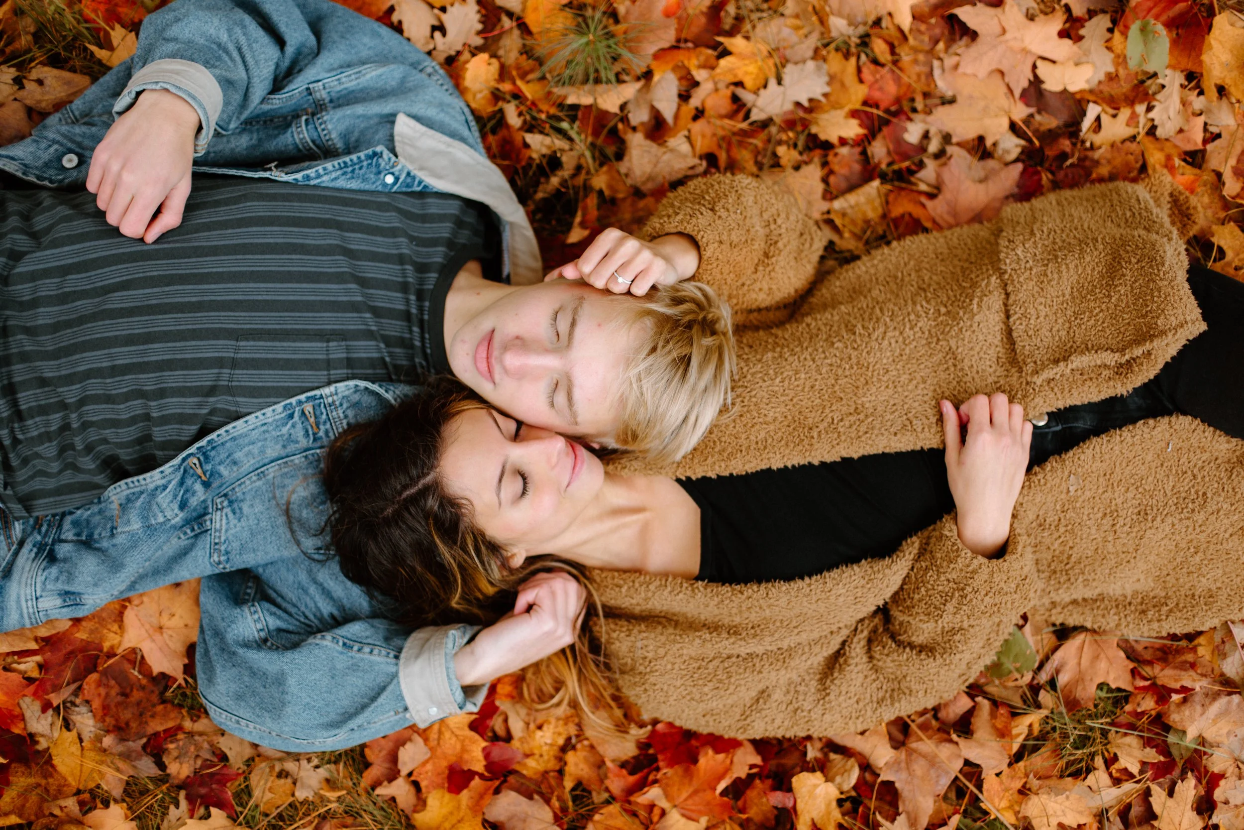 Three young women lying on a bed of autumn leaves outdoors, appearing relaxed and happy with their eyes closed.