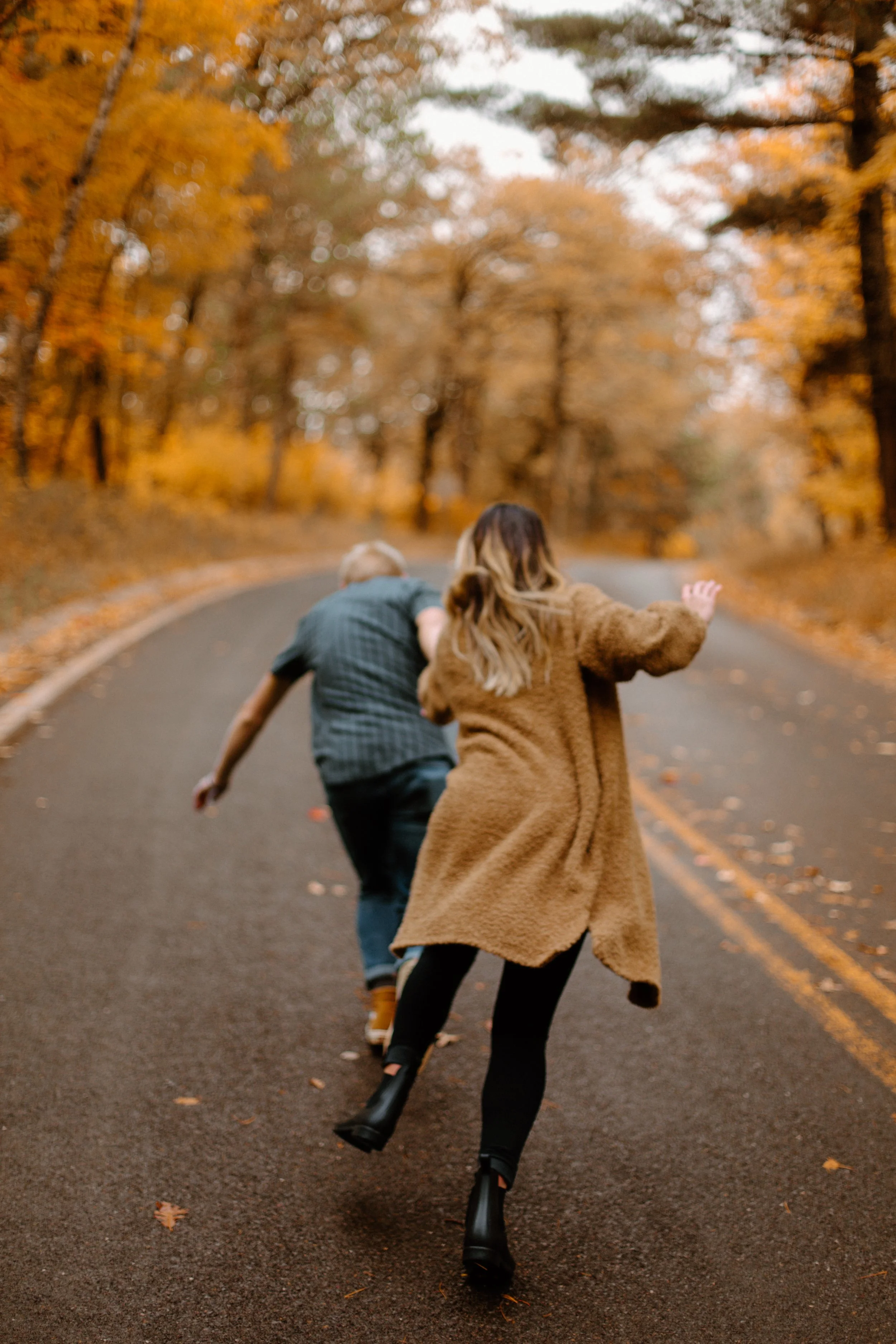 Two people running on a winding road through a forest with autumn leaves, one wearing a brown coat and the other in a plaid shirt.