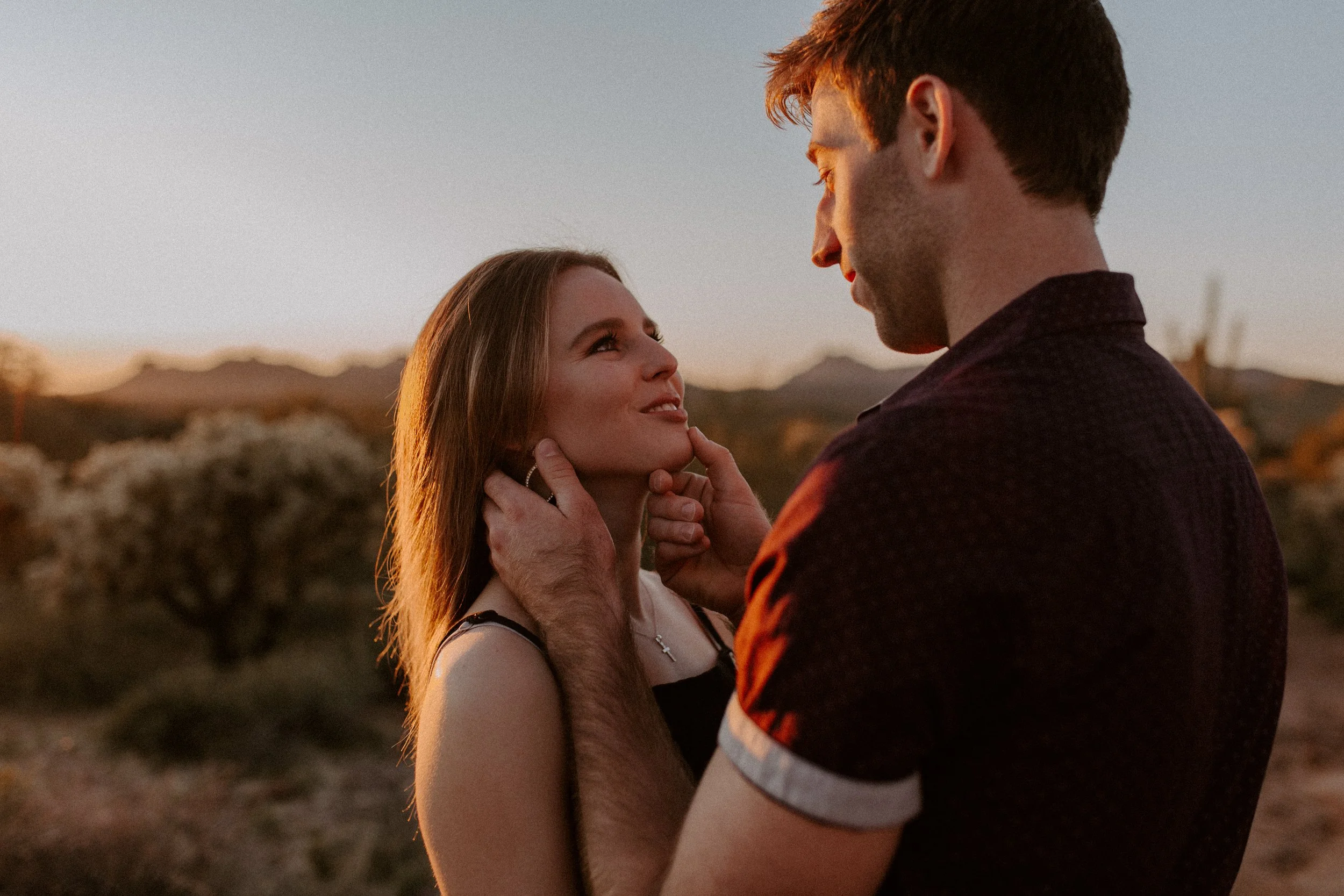 A man and woman in an outdoor setting during sunset, looking at each other with affection. The woman has long hair and is wearing a cross necklace, while the man has short hair and is wearing a dark shirt. The woman is gently touching the man's face,