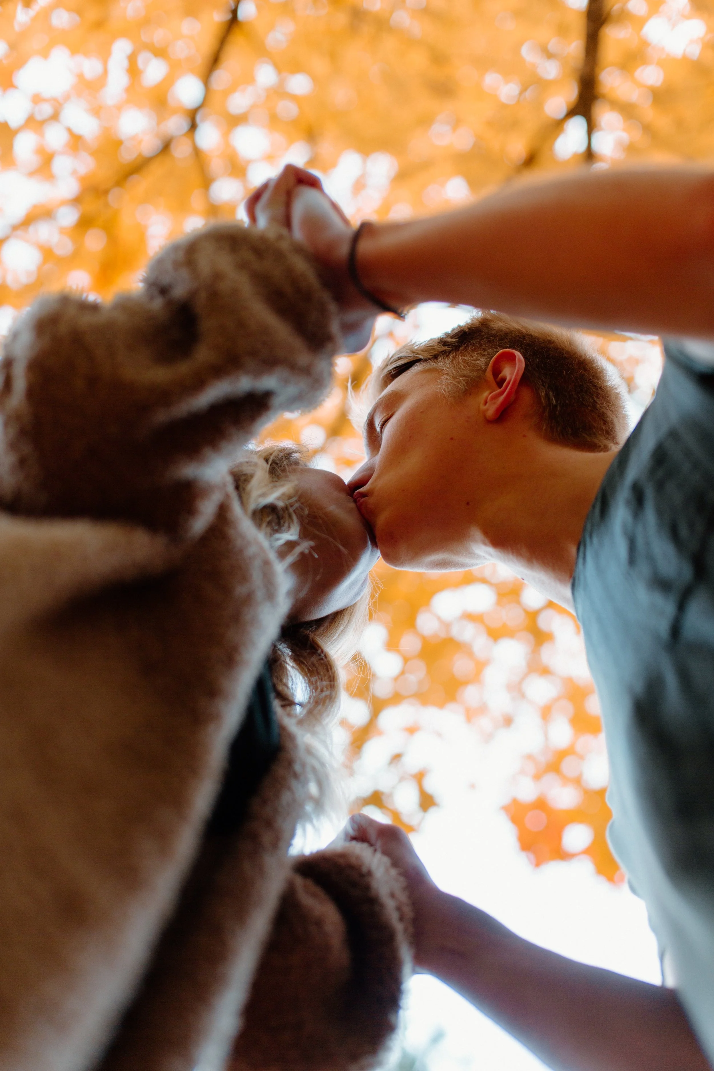 A young man and woman sharing a kiss outdoors during autumn, viewed from below with leaves and sky in the background.