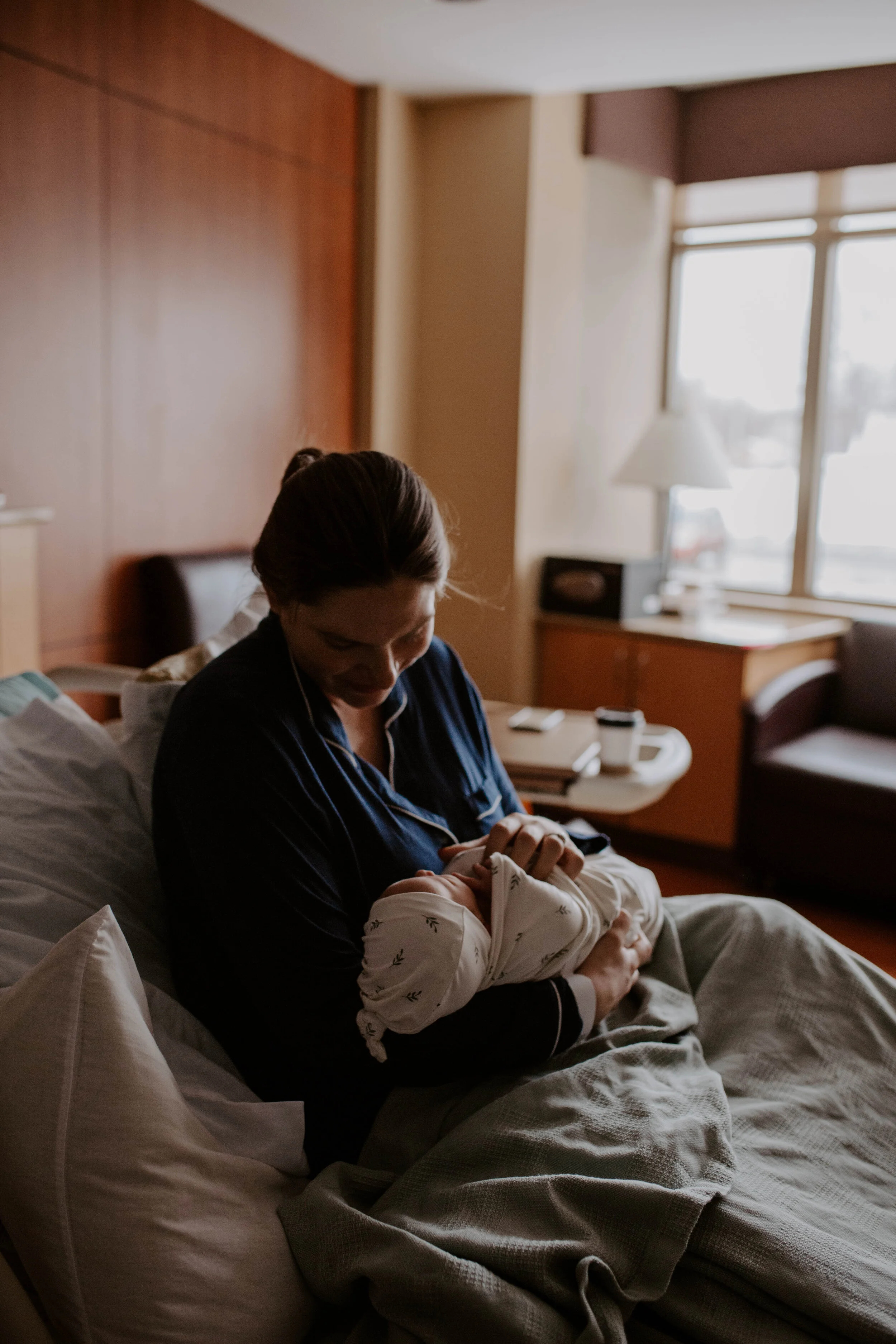 A woman sitting on a hotel bed holding a baby in her arms, with a warm sunlight coming through the window in the background.