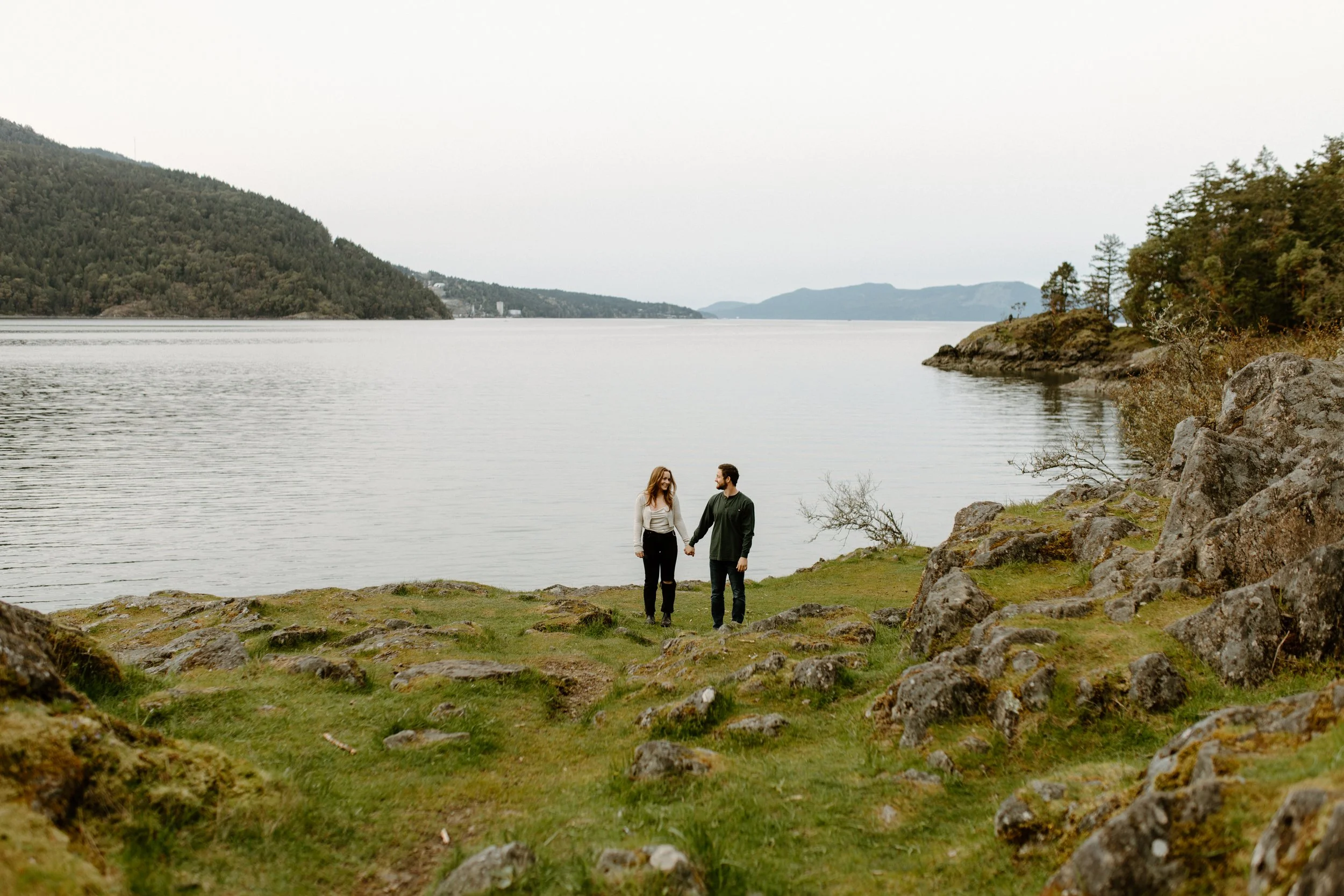 A couple holding hands and walking by a lake with forested hills and mountains in the background.