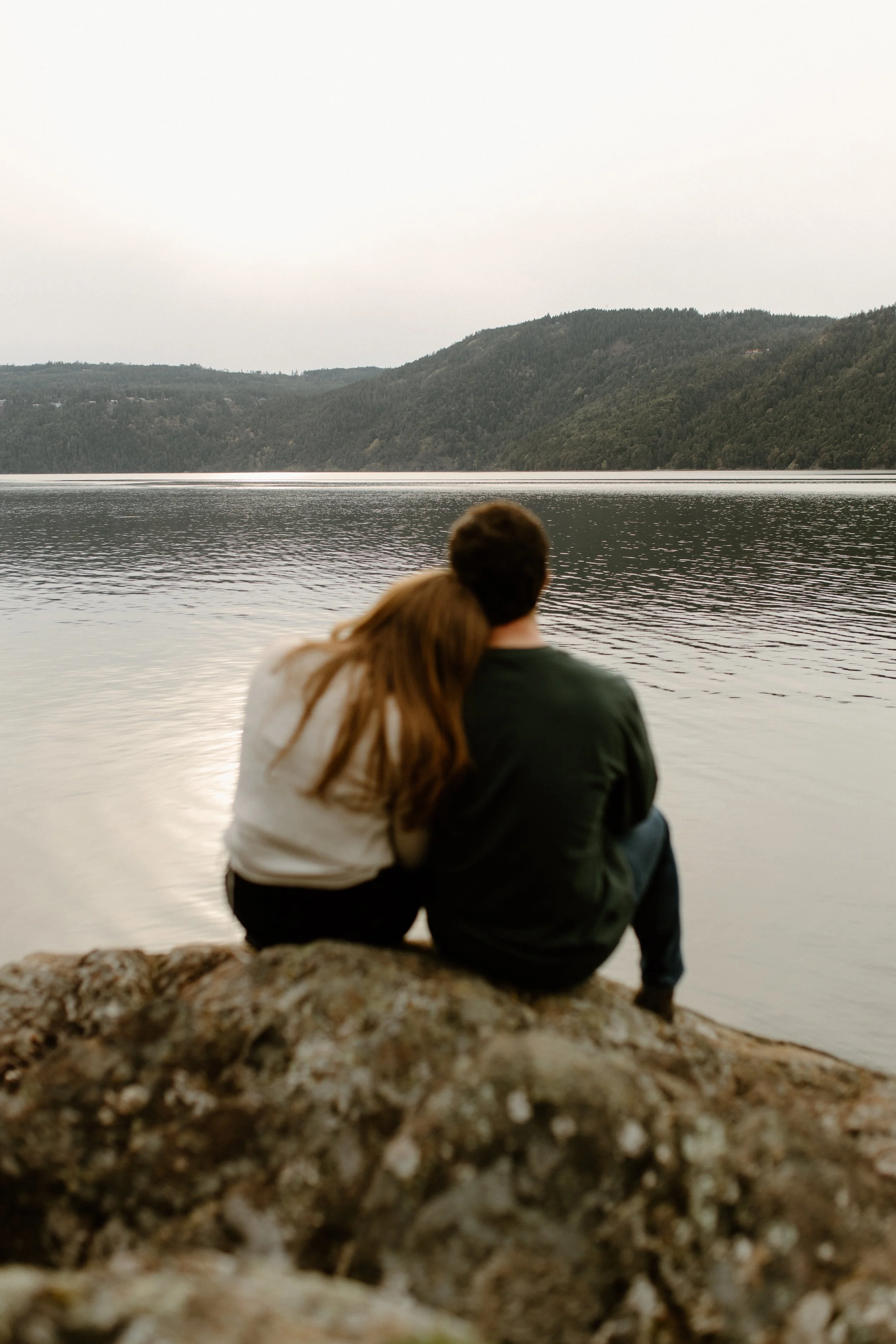 A couple sitting on a rock by a lake, gazing at the distant hills covered in trees under an overcast sky.