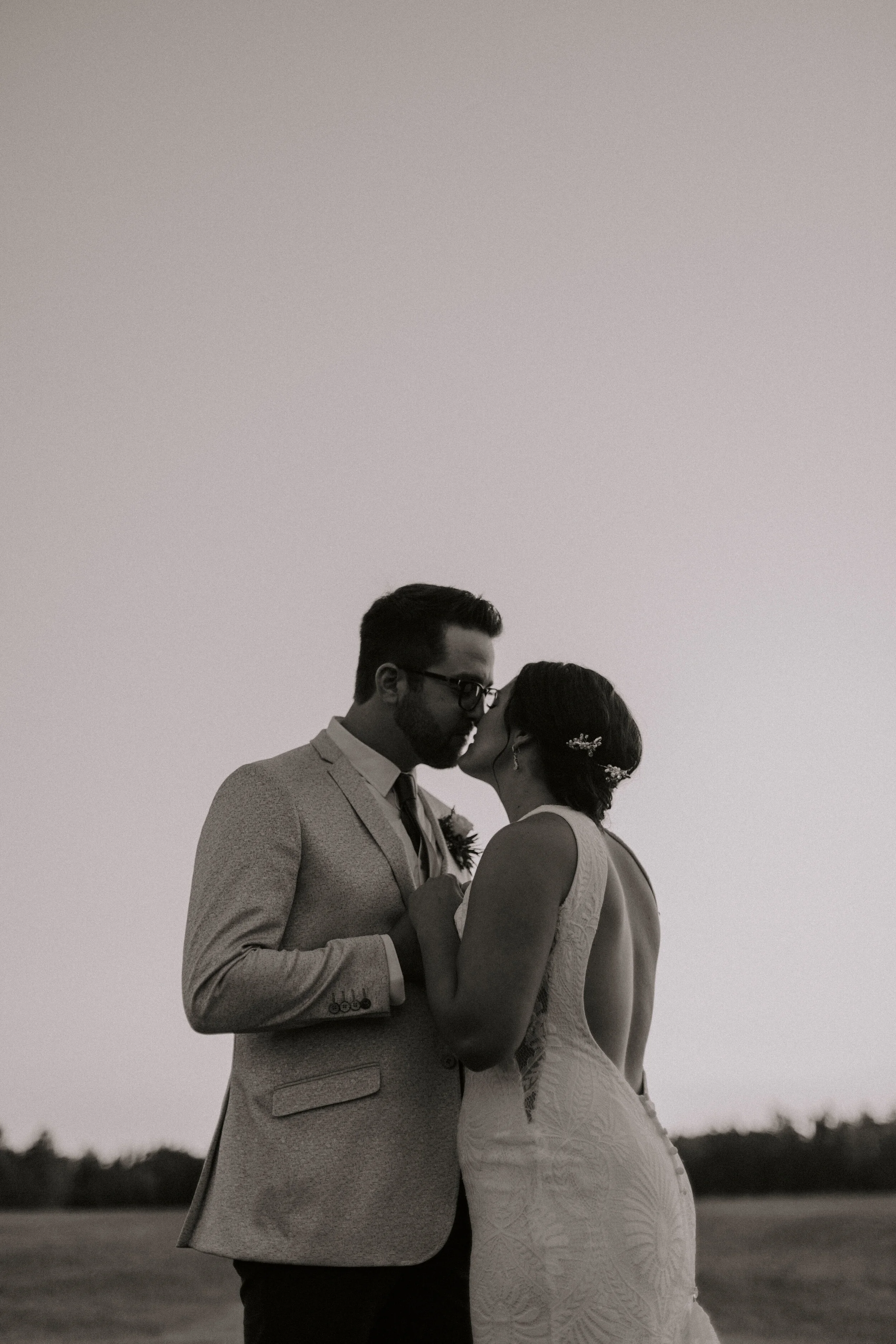 A black-and-white photograph of a bride and groom kissing outdoors during sunset. The groom is wearing glasses and a light suit, and the bride is in a lace wedding dress with a floral hair accessory. They are holding hands, and the background feature