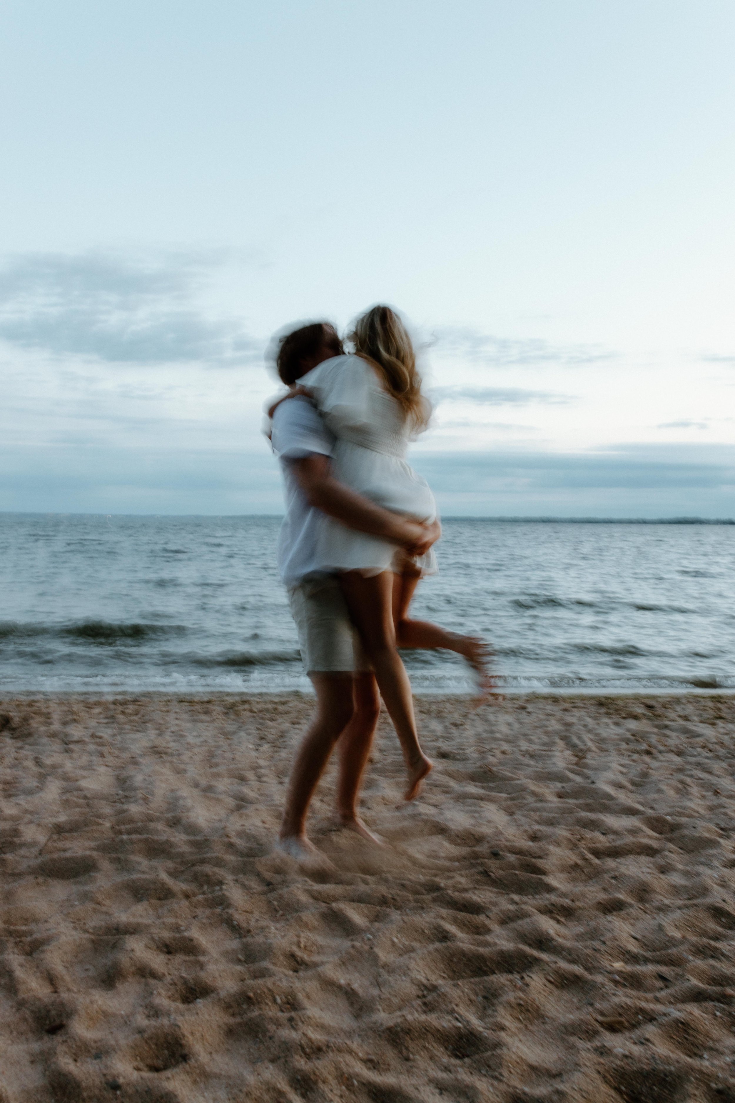 A couple on a beach, with the man lifting the woman in his arms, both smiling, with the ocean and sky in the background during sunset.