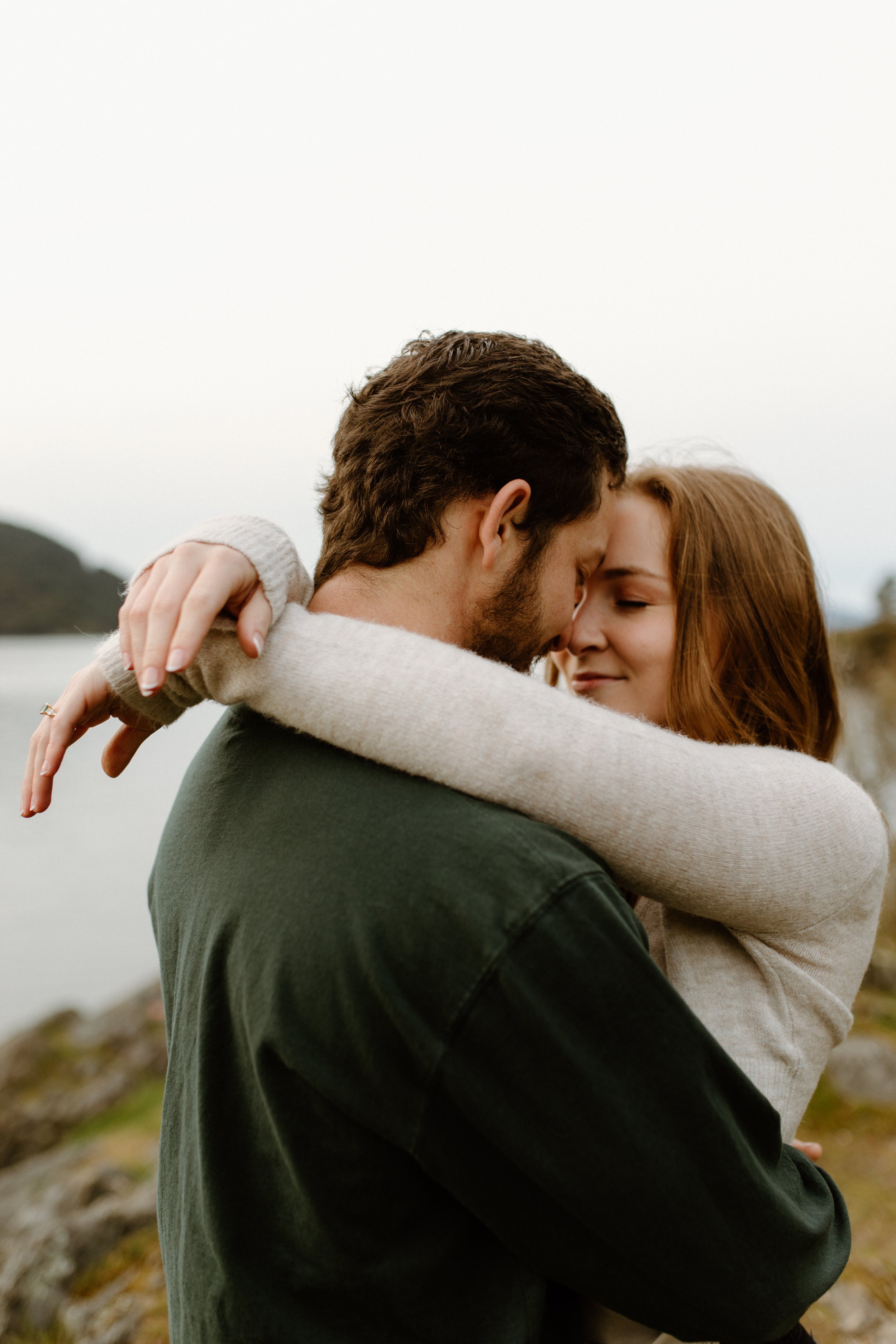 A couple embracing outdoors near a body of water, with the man facing away and the woman with her face close to his, eyes closed, both with content expressions.