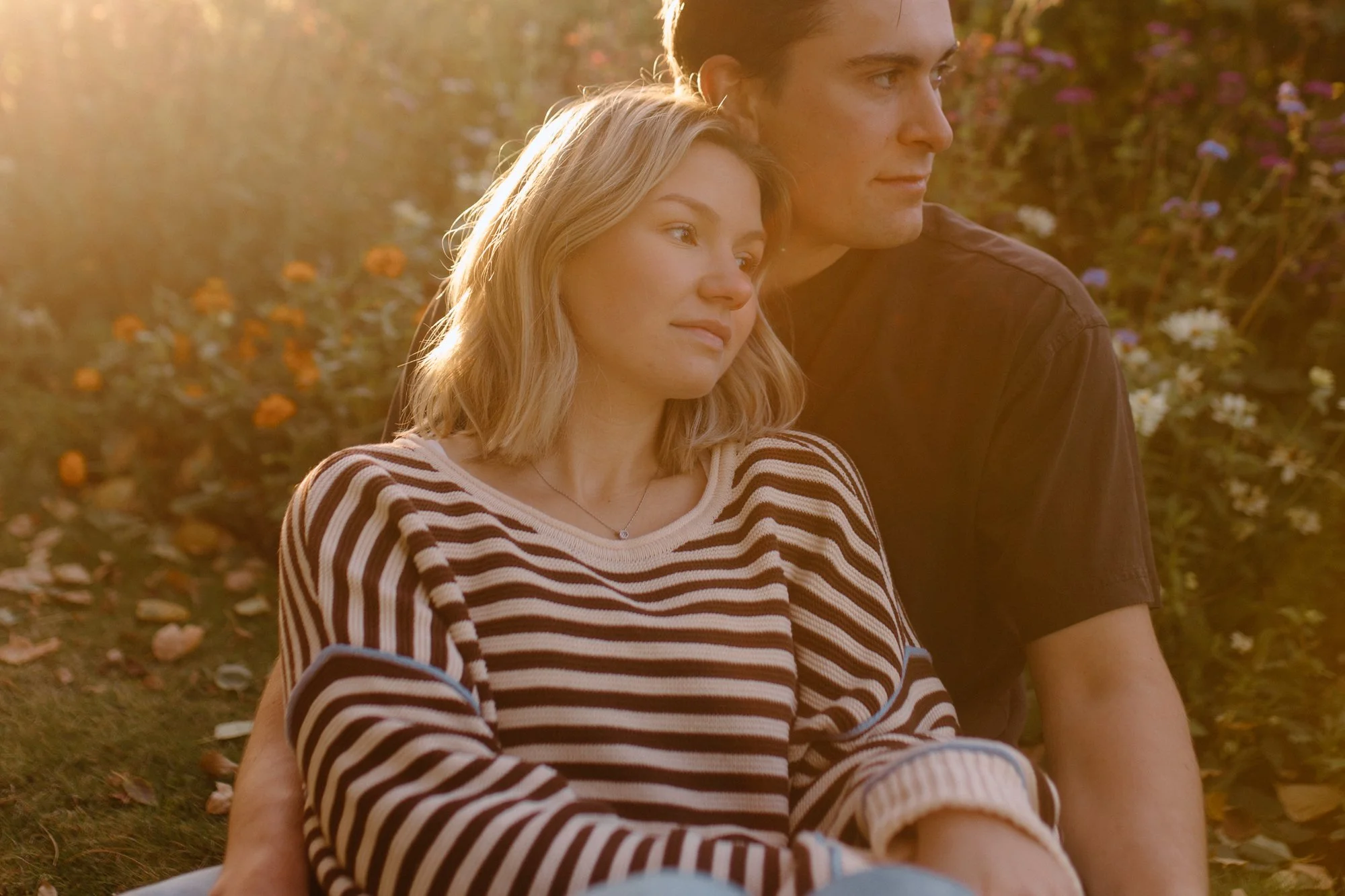 A young woman with blonde hair and a striped shirt sitting next to a man with dark hair, both enjoying a moment together outdoors in a garden during sunset.