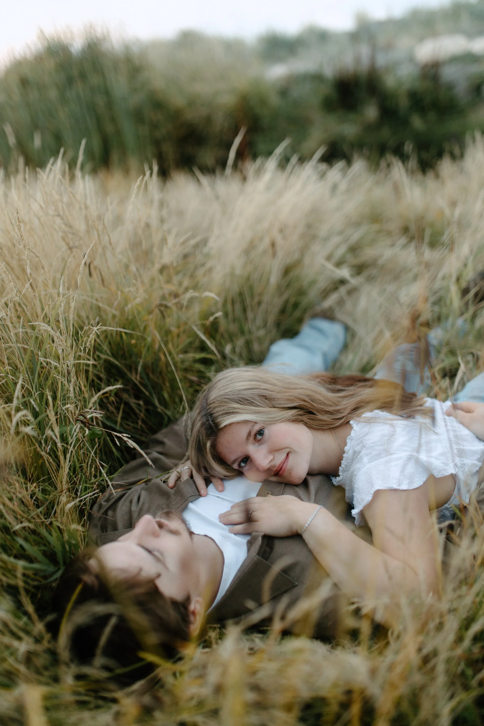 A young woman with blonde hair lying in grass, smiling and looking at the camera, with two other people lying beside her in a field of tall grass.