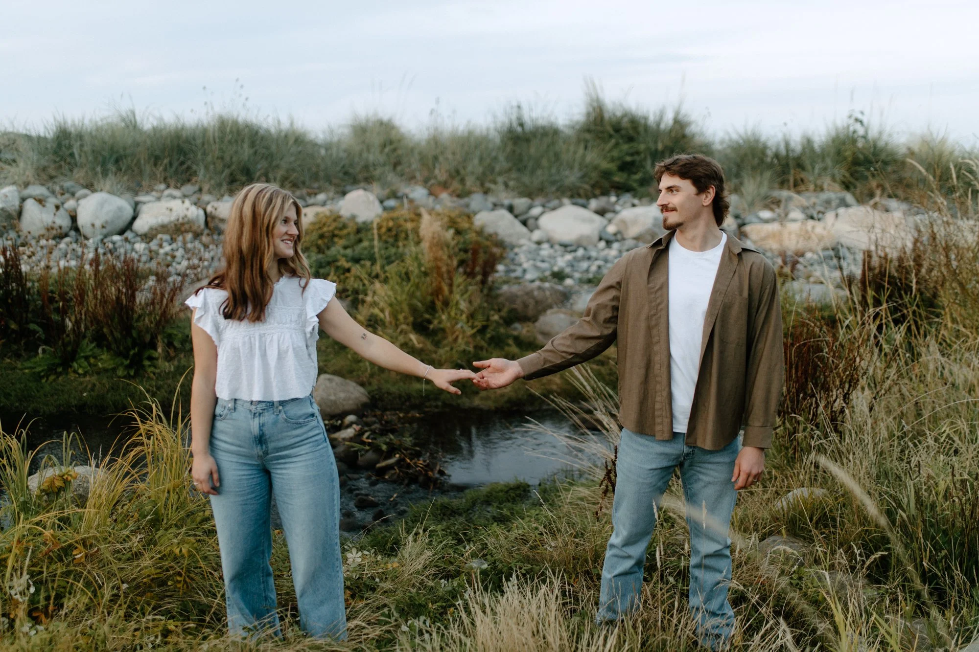 A young man and woman holding hands outdoors near a small stream, with rocks and tall grass, under a cloudy sky.