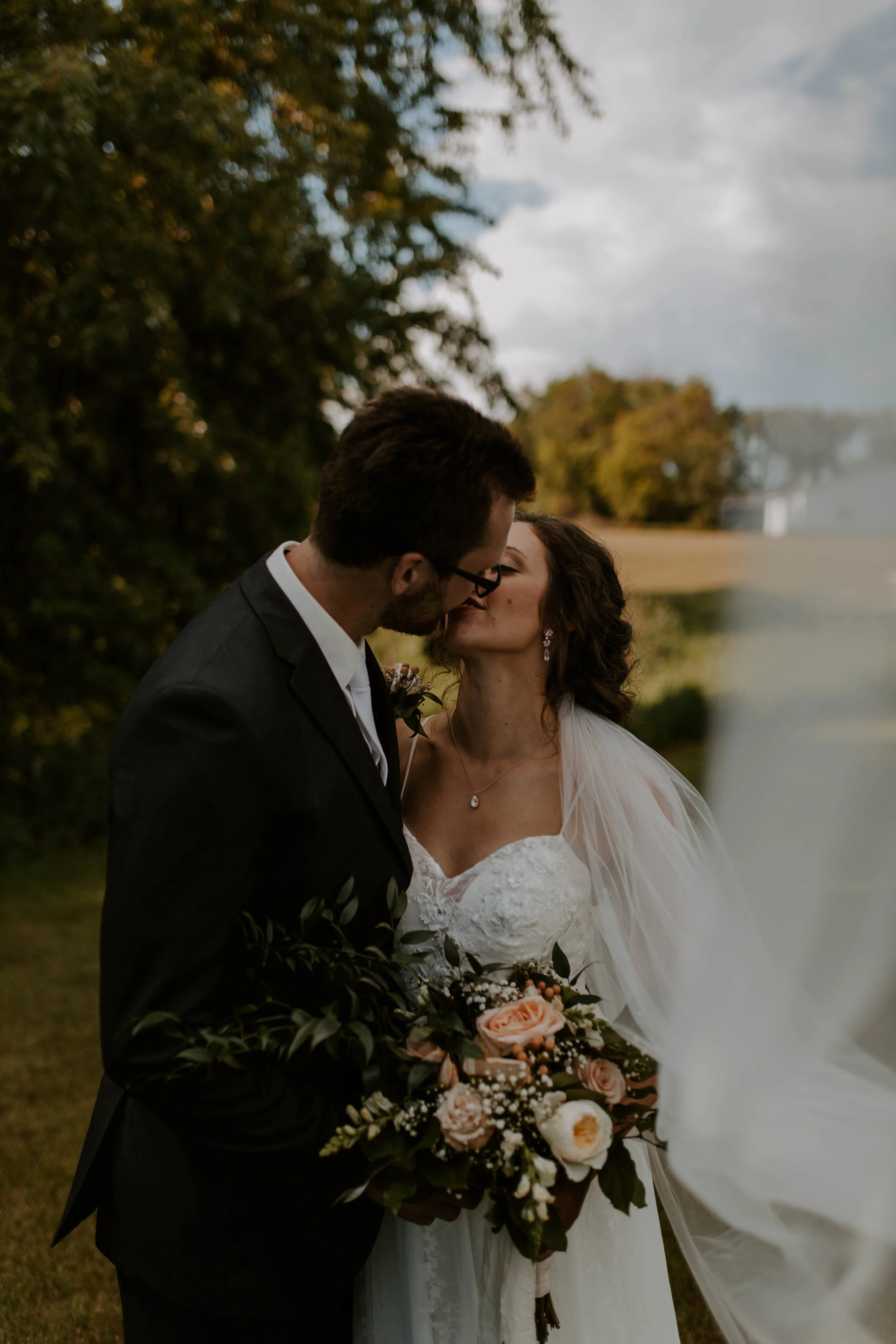 A bride and groom kiss at an outdoor wedding, with the bride holding a bouquet of roses and greenery; trees and a cloudy sky are in the background.