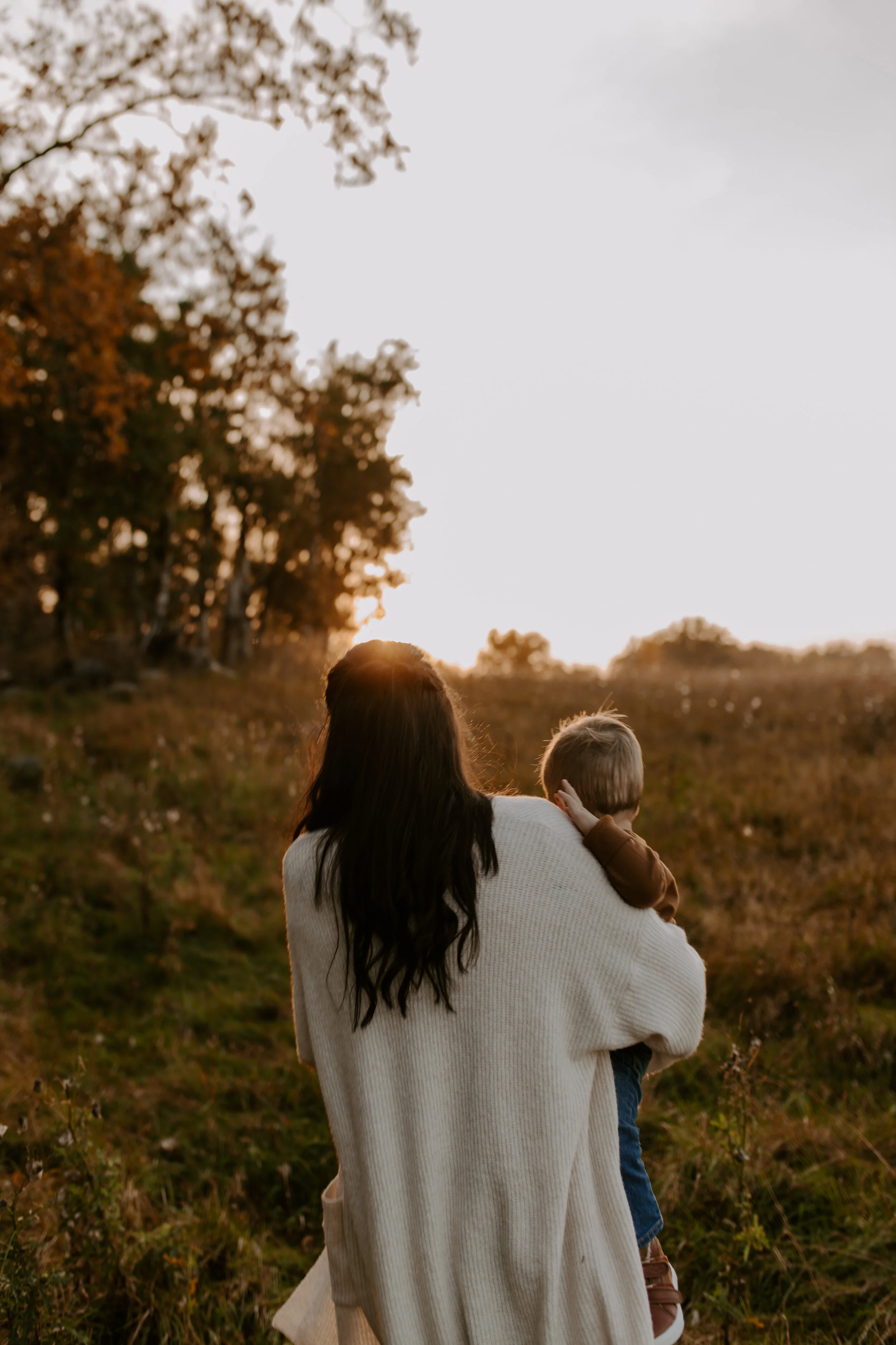 A woman holding a young boy in a rural outdoor setting during sunset, with trees and open field in the background.