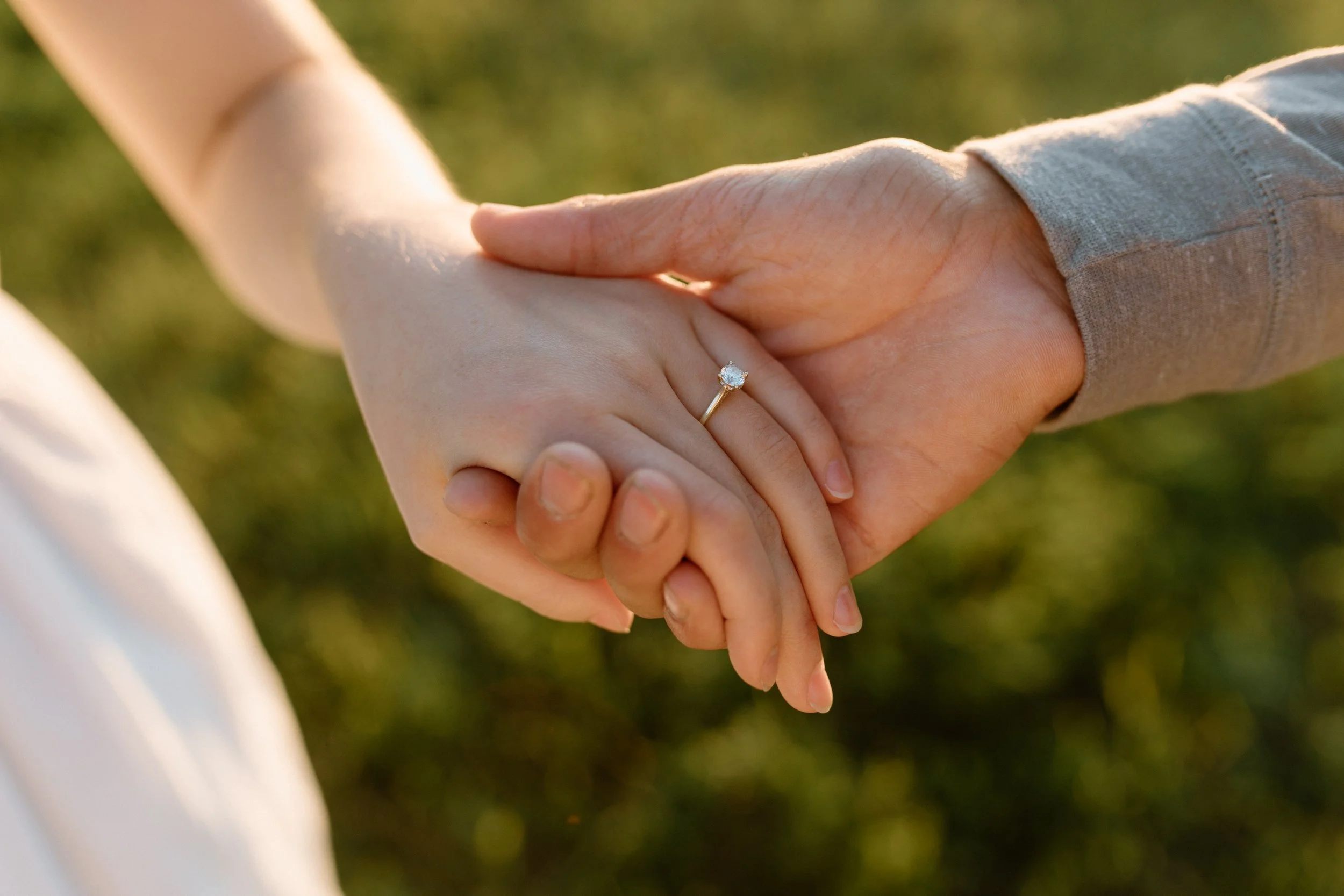 A close-up of a hand wearing a wedding or engagement ring, holding another person's hand, with a blurred green background.