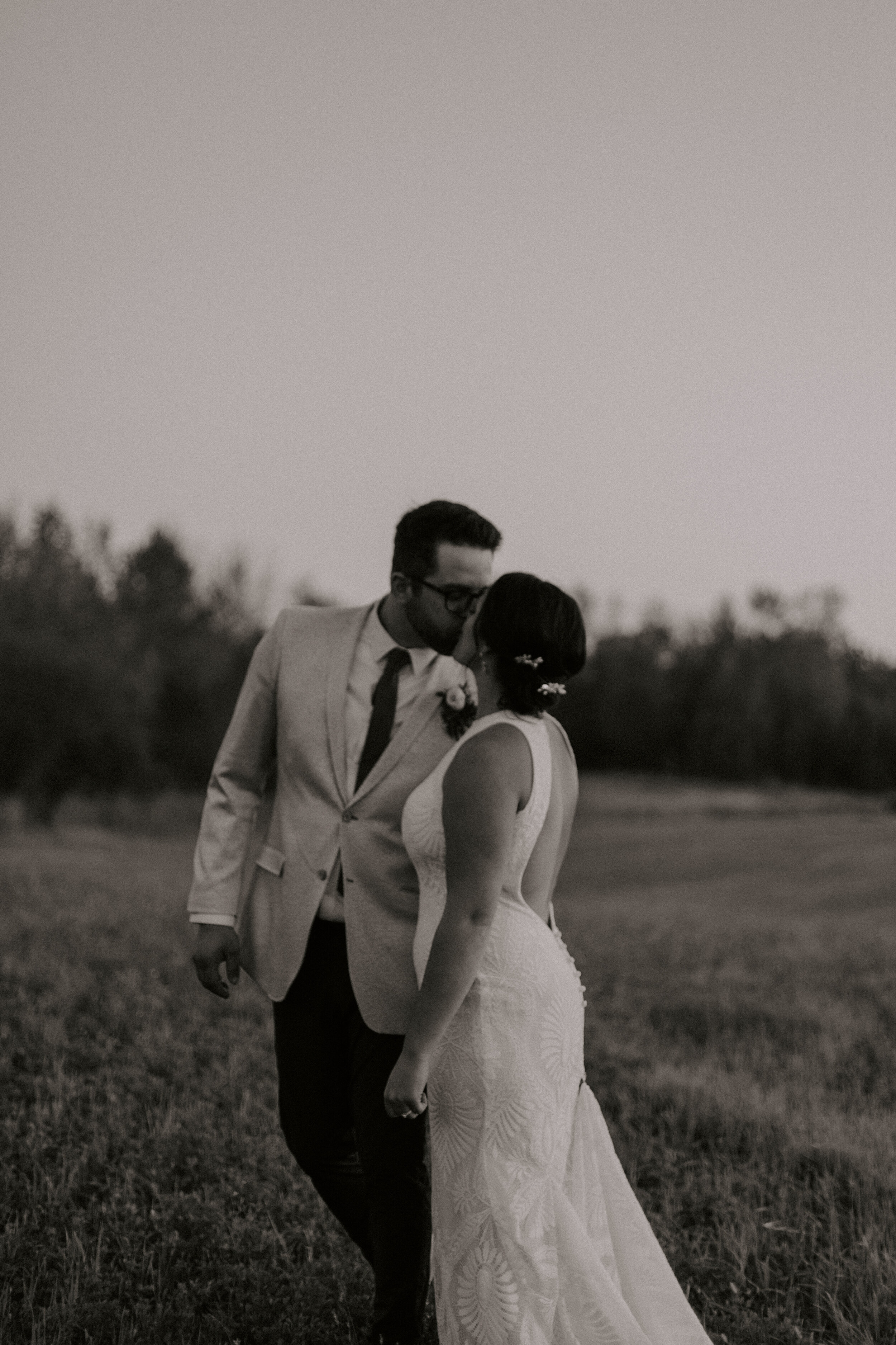 A black and white photo of a couple kissing outdoors, with the man in a suit and glasses and the woman in a white dress, standing on grass with trees in the distance.